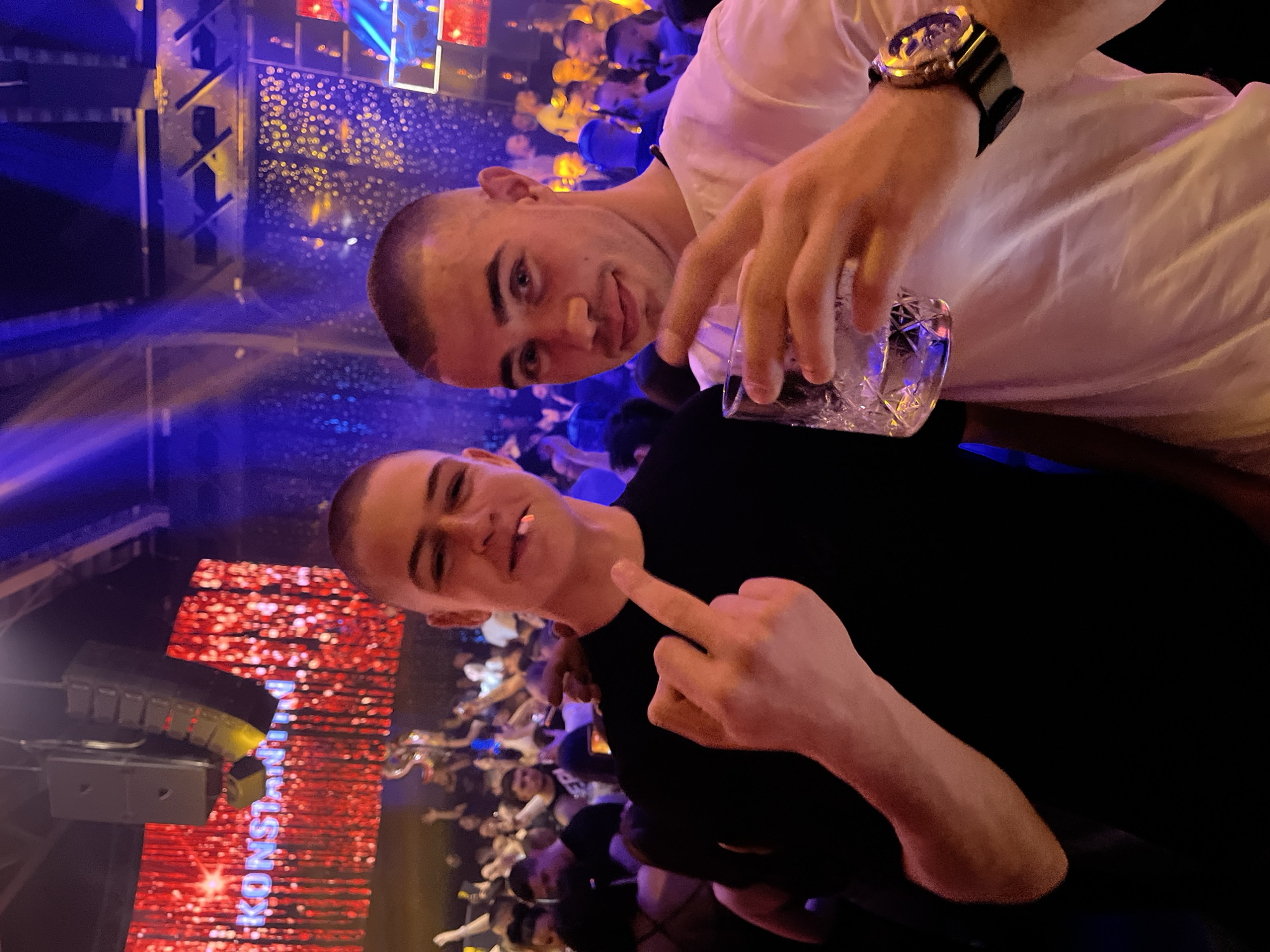 Two men are posing for a photo at a club in Burgas, Bulgaria. The man in the foreground is wearing a black shirt and giving the middle finger. The man behind him is wearing a white shirt and holding a glass of clear liquid. Both men have short hair. The club is crowded, and the background is brightly lit with blue and red lights. A screen with the word "KONSTANTIN" is visible behind them.