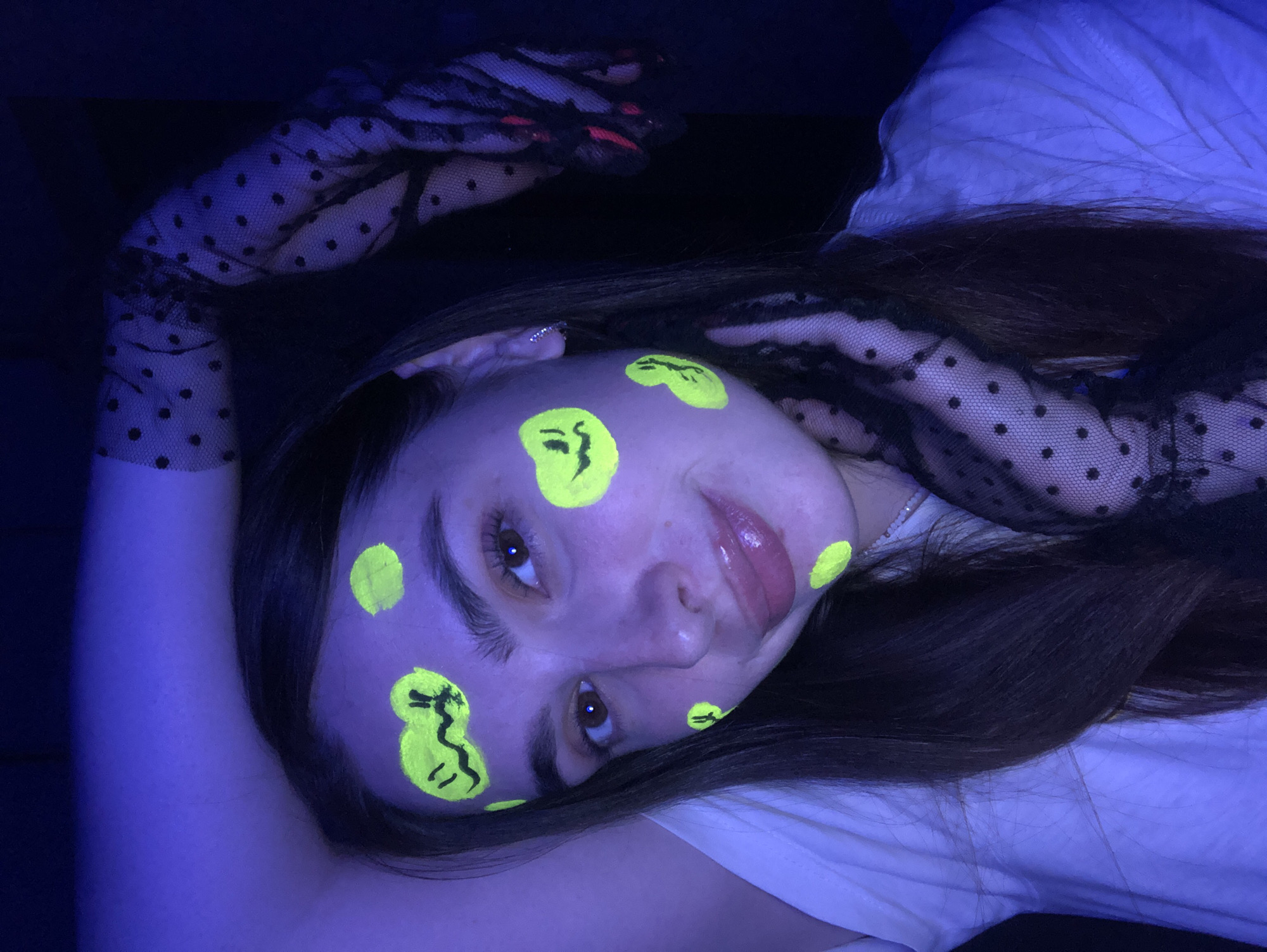 A young woman with long, dark hair is shown in a close-up, head-and-shoulders shot, posed in a dimly lit room under blue light. She is wearing a white t-shirt and black polka-dot gloves. Her face is adorned with neon yellow face paint that glows under the blacklight. The paint is applied in circular splotches, some of which are decorated with simple smiley faces. Her left arm is raised and bent behind her head, exposing the polka-dot glove. The background is dark and out of focus. There is no indication of the city of Noviy, Russia, in the image.