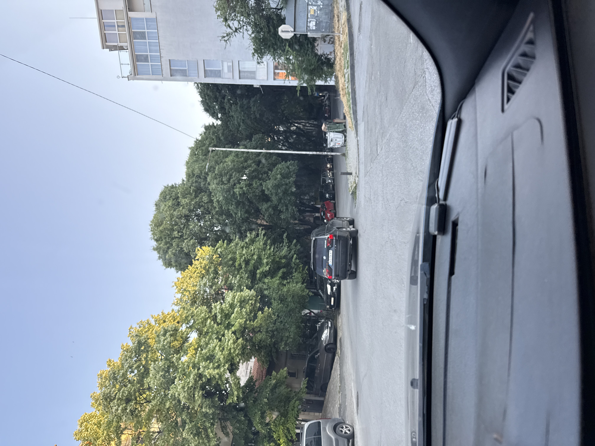 This image, captured from the interior of a vehicle looking forward through the windshield, depicts an urban street scene in Stara Zagora, Bulgaria. The location is confirmed by the "CH" district code visible on the license plate "CH 5156 AT" of a black SUV in the middle distance.
In the immediate foreground, the dark gray dashboard of the camera's vehicle is visible, including defroster vents and the base of the windshield.
The scene is dominated by a paved asphalt street with a slight curve to the right. On the left side of the street, a silver minivan or SUV is partially visible, while further back, a dark gray minivan is parked on an incline. Several other vehicles, including a red car, are visible parked or waiting on a side street to the left. In the center, a dark gray/black BMW X5 SUV is positioned at an intersection.
Mature trees with green and yellowish-green foliage line both sides of the street and fill much of the background, partially obscuring older buildings on the left, one with a tiled roof. On the right, a multi-story, light-colored building with multiple windows and balconies is prominent, extending into the upper right of the frame. A thin cable runs diagonally across the clear blue sky, originating from near the top of this building.
Near the intersection on the right, a tall, light-colored street lamp pole stands beside two green and white trash bins. Behind them, a round white regulatory sign is partially visible. No people are clearly discernible in the image. The lighting suggests late afternoon or early evening, with clear skies and direct sunlight illuminating the tops of the trees.