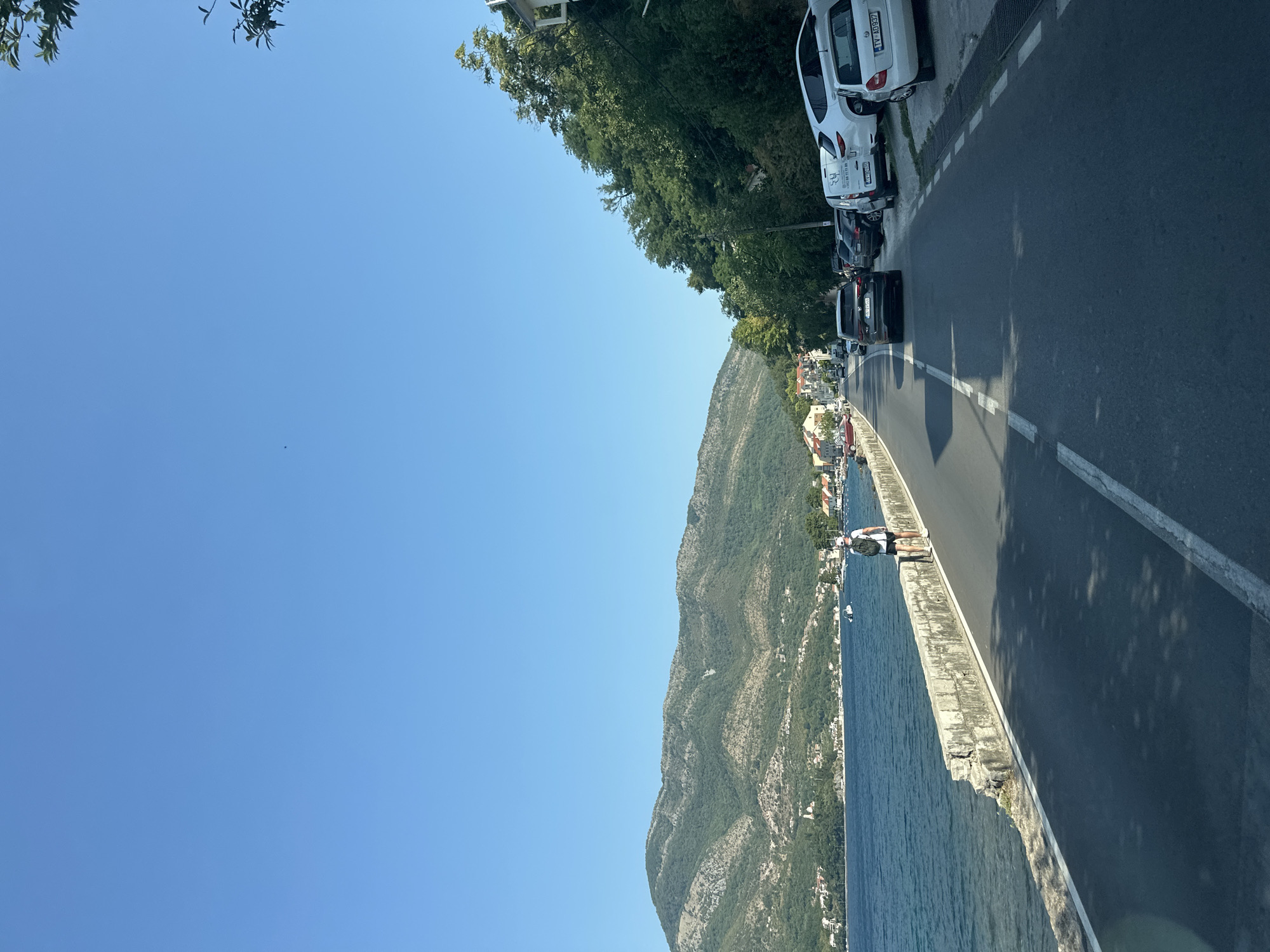 The image captures a coastal road in Donja Lastva, Montenegro, on a clear, sunny day. The road runs alongside a body of water, likely the Adriatic Sea, with a low stone wall separating the road from the water. In the distance, lush green mountains form a backdrop to the scene.
Several vehicles are parked on the right side of the road, including a white van and a white car with the license plate "TV AD937". A black SUV is driving away from the camera on the road. On the left side of the road, near the water, a person with a backpack is walking. A few boats are visible on the water. The coastline is dotted with buildings and trees, characteristic of a Mediterranean town. The sky is a vibrant, cloudless blue.