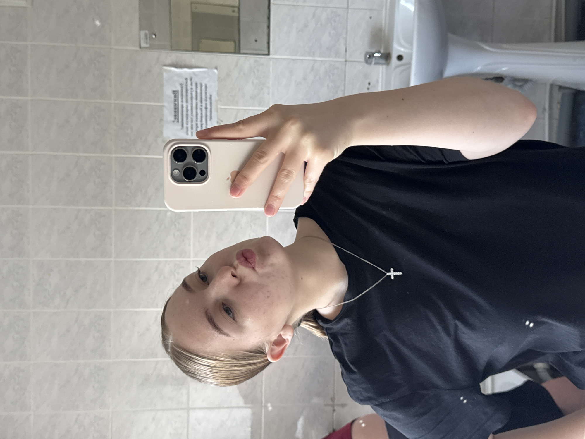 A young individual is captured in a mirror selfie taken in what appears to be a bathroom in Perm, Russia. The person, with fair skin and light-colored hair pulled back, is making a "kissing face" expression, looking towards the camera. They are wearing a dark, likely black, t-shirt and a silver chain with a small cross pendant around their neck.
The selfie is being taken with a light-colored (beige or pink) smartphone, likely an iPhone due to the distinct triple camera array and visible Apple logo on the back. The phone is held up, reflecting the person's hand and arm.
The background shows light-colored square tiles on the wall, characteristic of a bathroom. Partially visible on the right is a white ceramic sink or toilet bowl, reinforcing the bathroom setting. Above the sink, a metal faucet or fixture is visible. A crumpled piece of white paper with indistinct text, appearing to be in Cyrillic script, is taped to the mirror or wall behind the person. The lighting suggests an indoor environment. The overall mood conveyed is casual and playful.