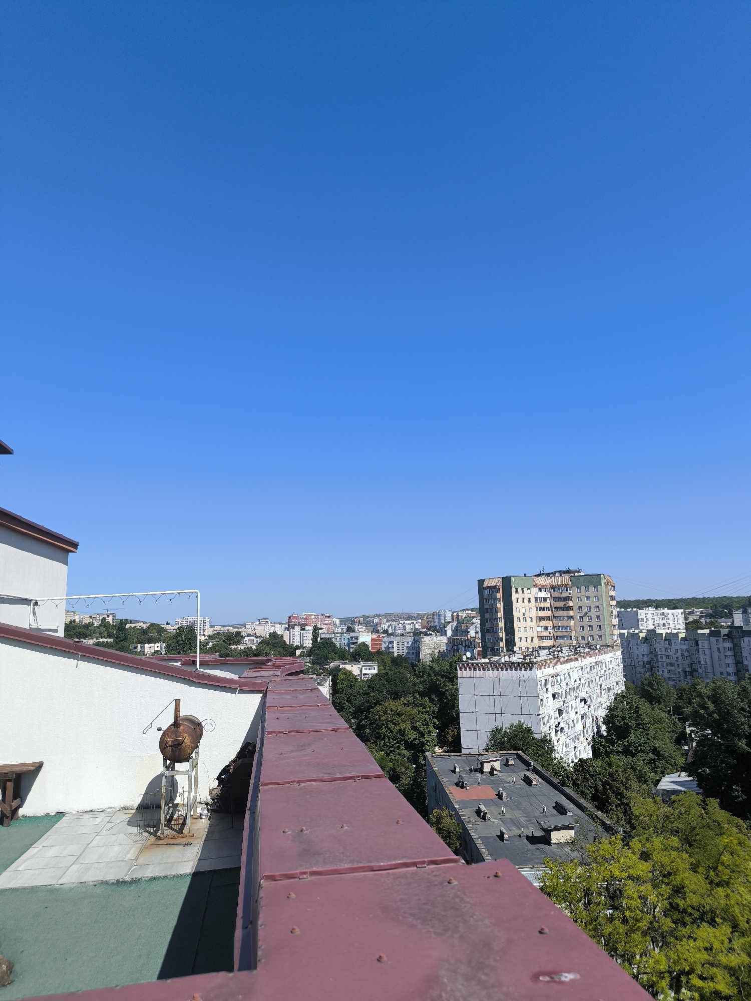 This is a high-angle, vertical, outdoor photograph taken on a sunny day with a clear blue sky dominating the upper portion of the frame. In the foreground, a burgundy-colored metal roof edge with visible rivets runs horizontally across the bottom of the image. To the left, a white building wall with a maroon-colored roofline is visible, with a wooden bench and a rusty barbeque grill on a tiled surface in front of it.
The middle ground of the image reveals a city skyline populated with apartment buildings of varying heights and architectural styles, interspersed with lush green trees. One prominent, tall apartment building with a green top is centrally located in this section. To the right, more buildings and trees extend towards the horizon. The background consists of distant buildings and a hazy horizon line.
Based on architectural styles and the presence of similar residential buildings, this scene is highly characteristic of the city of Chișinău, Moldova, specifically resembling views from residential areas within the city. No people are visible in the image.