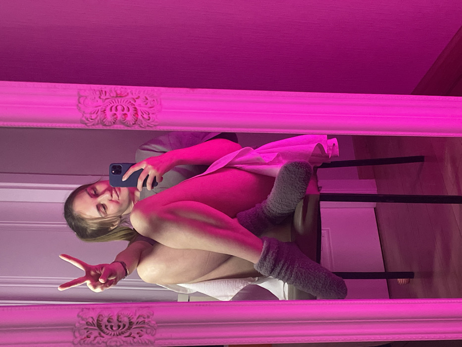 A woman is reflected in a mirror, sitting on a chair. She is wearing a short, light-colored dress and fuzzy socks. She holds a blue phone in one hand, presumably taking the picture. With the other hand, she makes a peace sign. The image has a pinkish hue, likely due to lighting. The mirror has a white, ornate frame. Parts of a door and a chair are also visible.