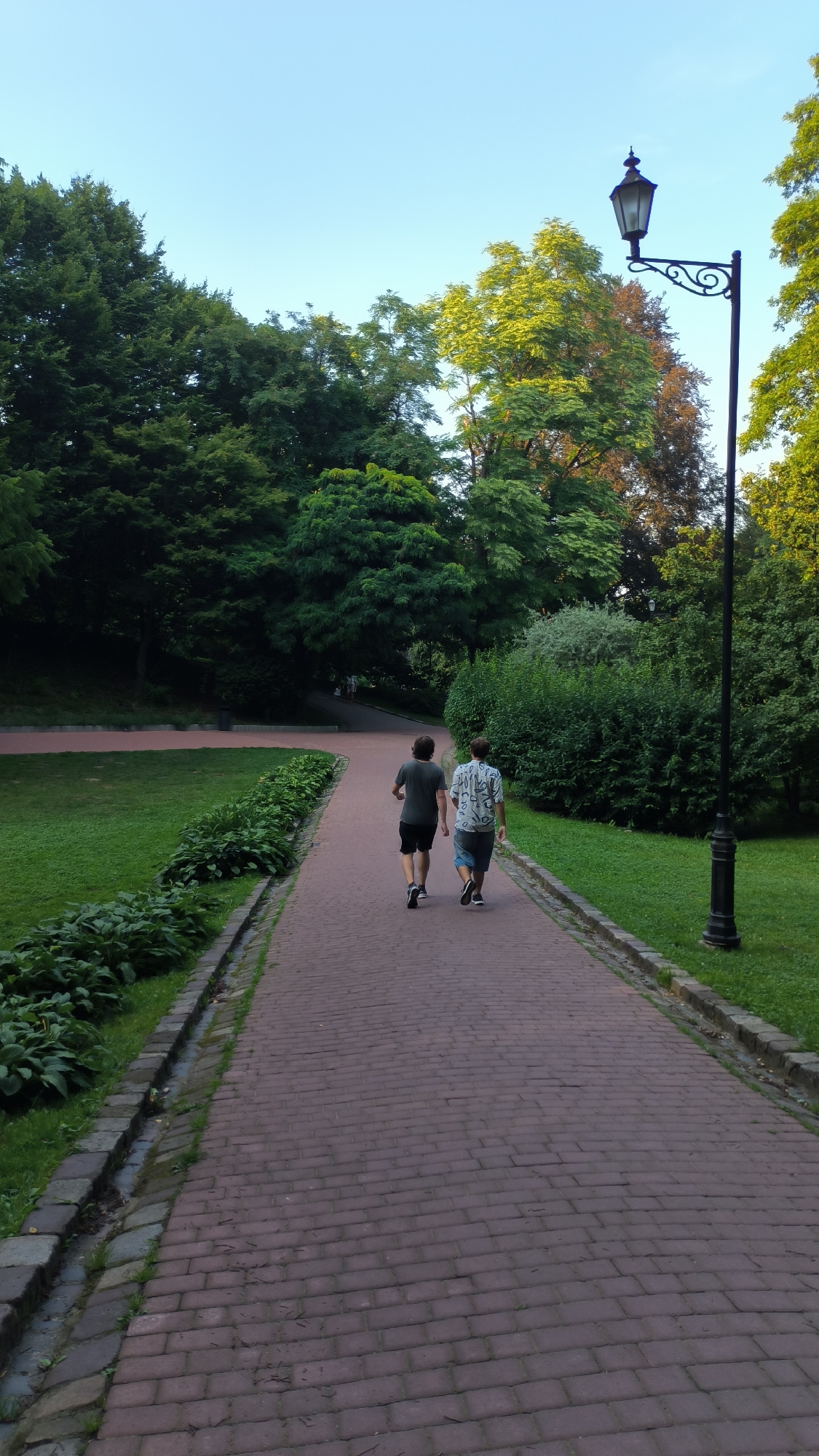 Two individuals are walking away from the camera on a brick-paved pathway in a park. The person on the left is wearing a dark t-shirt and black shorts, while the person on the right is wearing a patterned short-sleeved shirt and blue shorts. They are walking amidst lush green trees and grass under a clear sky. A traditional lamppost stands to the right of the pathway. This scene appears to be located in Stryiskyi Park in Lviv, Ukraine, a renowned urban green space known for its historical significance and diverse flora. The park's landscape, with its winding paths, manicured lawns, and mature trees, is consistent with the visual cues in the image.