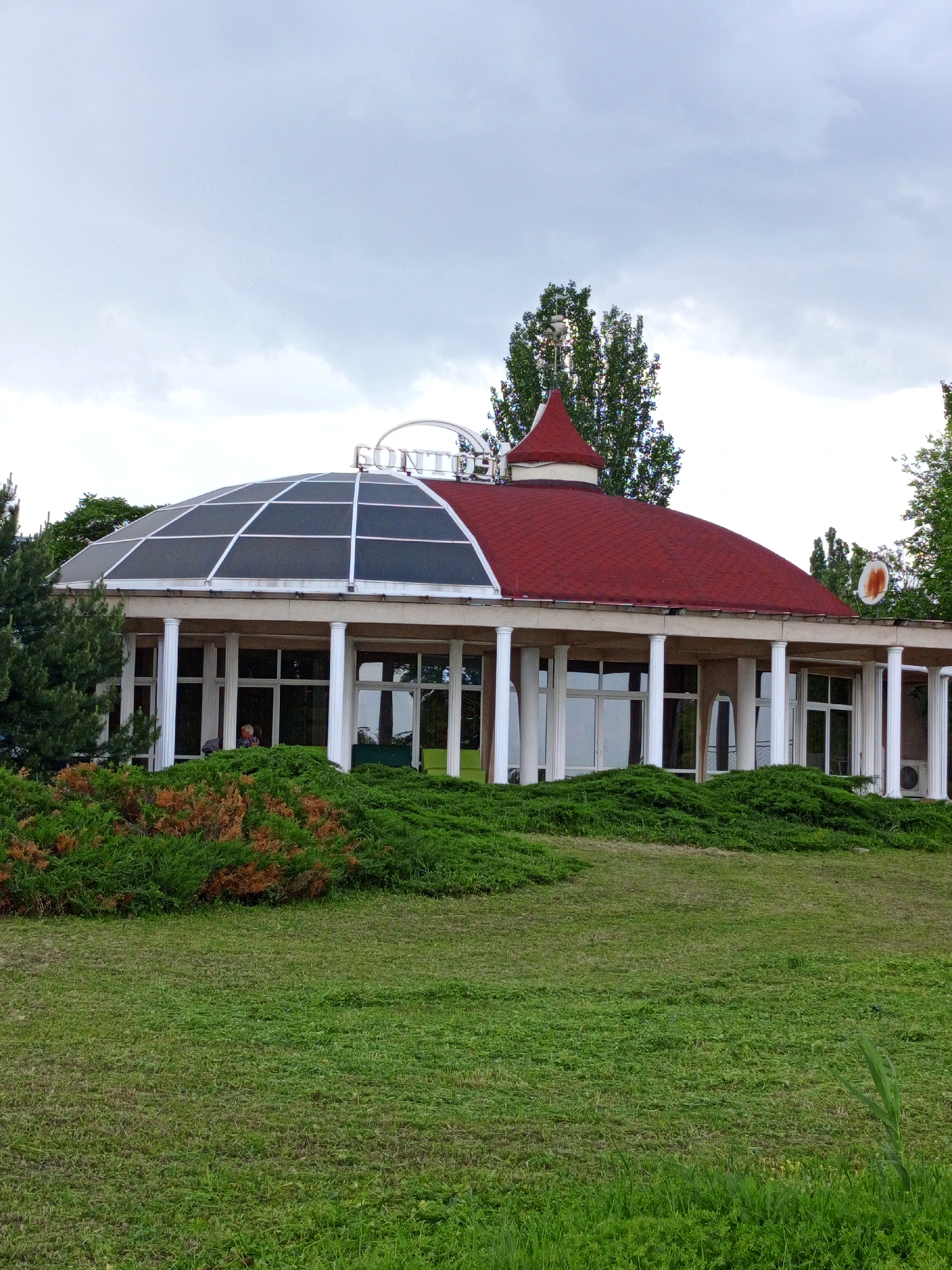 The image shows a one-story building in an urban setting. The building has a unique architectural design, featuring a red-tiled roof on one half and a glass-paneled dome on the other. White pillars support the structure, and large windows are visible beneath the roofline. The building is surrounded by greenery, including a manicured lawn and bushes. The sky is overcast, suggesting a cloudy day. A sign reading "SONTO" is visible on the building. A satellite dish is attached to the building near the red tiled roof. An air conditioning unit sits next to one of the pillars. The location is the city of Unknown, Unknown.
