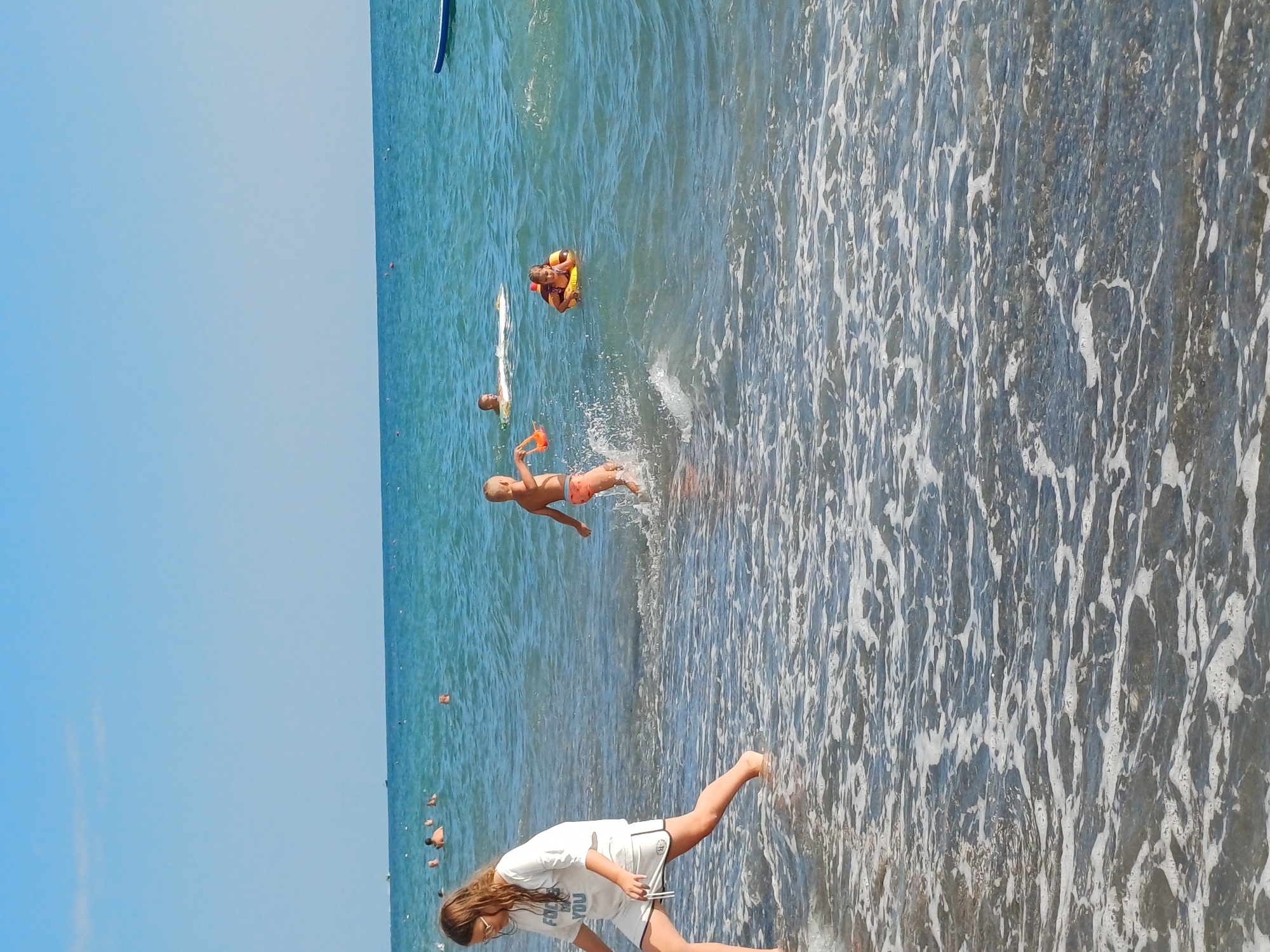 The image shows a beach scene in Sochi, Russia. The main subjects are people enjoying the water. In the foreground, a young girl with long brown hair and glasses, wearing a white t-shirt and shorts, is standing in shallow water. She appears to be looking down at something. Further out in the water, a boy in orange swim trunks is mid-air, seemingly jumping or diving, with a splash around him. Another person, wearing a yellow floatation device, is visible further back in the water.
The scene is set at the sea with clear blue sky above and the ocean with gentle waves. The water is a mix of deep blue and lighter, foamy areas near the shore. The time of day appears to be daytime, with bright sunlight illuminating the scene. The overall mood seems relaxed and joyful, typical of a beach holiday.
On the girl's t-shirt, there is some visible text, partially obscured, which appears to read "FOOD FOR YOU". The location cues of the clear blue sky and the general appearance of a popular beach resort align with the context of Sochi, Russia.