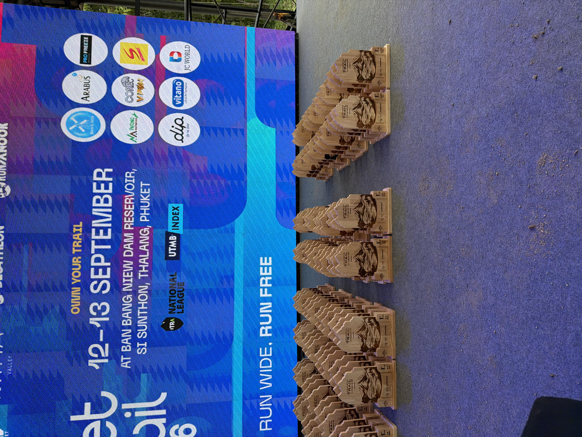 This image shows a large digital screen displaying event information and sponsorship logos, positioned behind rows of wooden trophies. The screen text announces "OWN YOUR TRAIL 12-13 SEPTEMBER AT BAN BANG NIEW DAM RESERVOIR, SI SUNTHON, THALANG, PHUKET." Below this text are several circular logos representing sponsors such as "Duchy Miu," "ARABUS," "PRO FREEZE," "MA PATONG," "CORE Vison," "S CHINOS," "dip," "vitano," and "JC WORLD." There are also smaller logos for "ITRA NATIONAL LEAGUE" and "UTMB INDEX." The trophies, arranged in neat rows on a stage, are shaped like mountains with a banner across the top indicating "PHUKET TRAIL 12-13 September 2020." Each trophy has a base with sponsor logos and information, including "1st PLACE." The foreground shows a blue carpeted surface with some scattered dirt or dust. The background, visible through the screen, suggests an outdoor environment with trees.