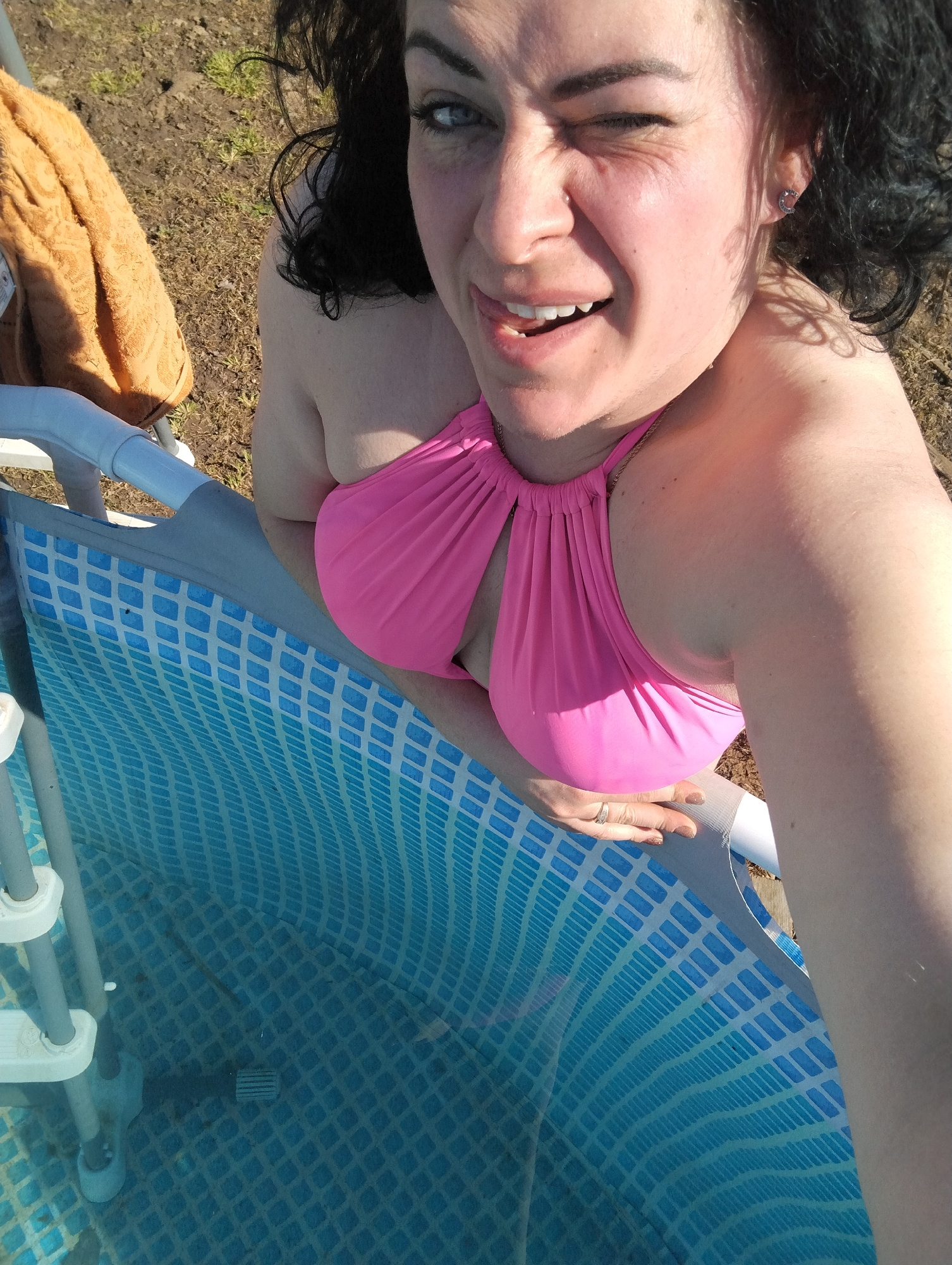 A woman with dark curly hair, wearing a pink swimsuit, winks at the camera while standing next to a blue and white above-ground pool. A yellow towel is draped over the pool's edge in the background. The ground appears to be brown and grassy. The photograph was likely taken outdoors during daylight. There is no specific information to confirm the location as Grishanki, Russia.