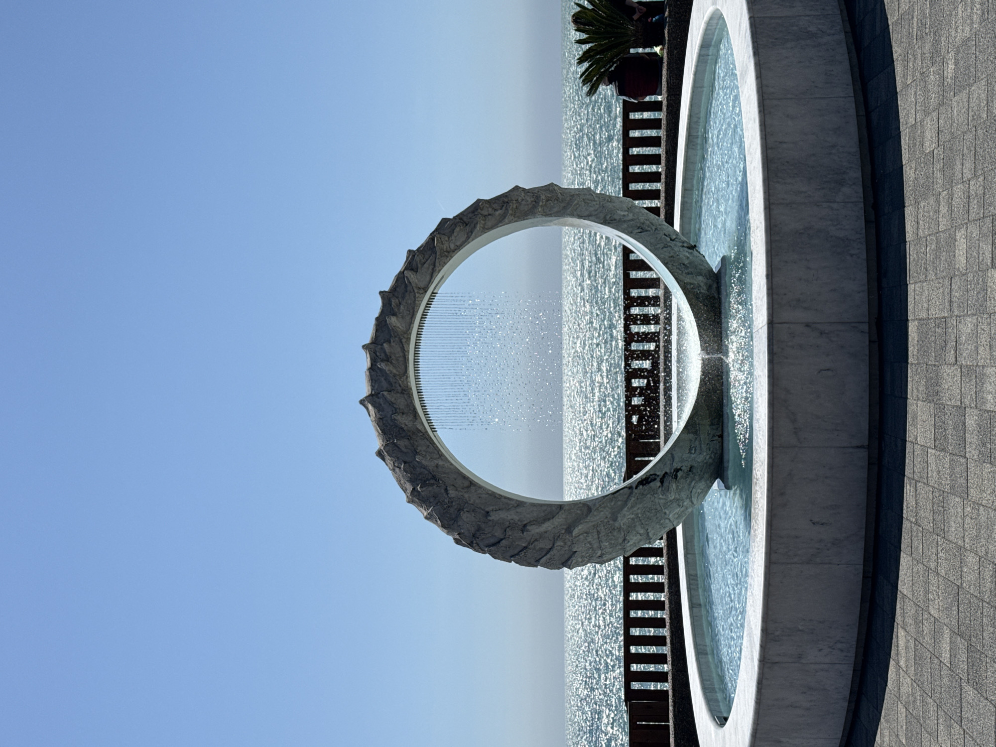 An outdoor scene in Sochi, Russia, features a large, striking modern water fountain under a clear blue sky. The central element of the fountain is a substantial circular archway, crafted from what appears to be rough, textured grey stone, with jagged protrusions along its outer edge. Within the arch, numerous fine streams of water descend, creating a delicate, shimmering water curtain that falls into a circular basin below.
Beyond the fountain, a vast expanse of the sea sparkles intensely under bright sunlight, indicating a clear, sunny day. Between the fountain and the glistening sea, there is a dark-colored structure, possibly a pier or promenade, with prominent wooden slatted railings or benches. In the upper right background, behind this structure, the fronds of a palm tree are visible, along with glimpses of indistinct figures, suggesting people are present, perhaps sitting or walking along the waterfront.
To the right of the fountain, a wide, paved area extends, made of light-colored, rectangular tiles, possibly granite or marble, contributing to the modern aesthetic of the space. A darker, textured wall or architectural feature is visible further to the right. The overall setting appears to be a well-maintained public park or promenade along the coastline, offering scenic views of the sea and an elegant water feature. No visible text is present in the image.