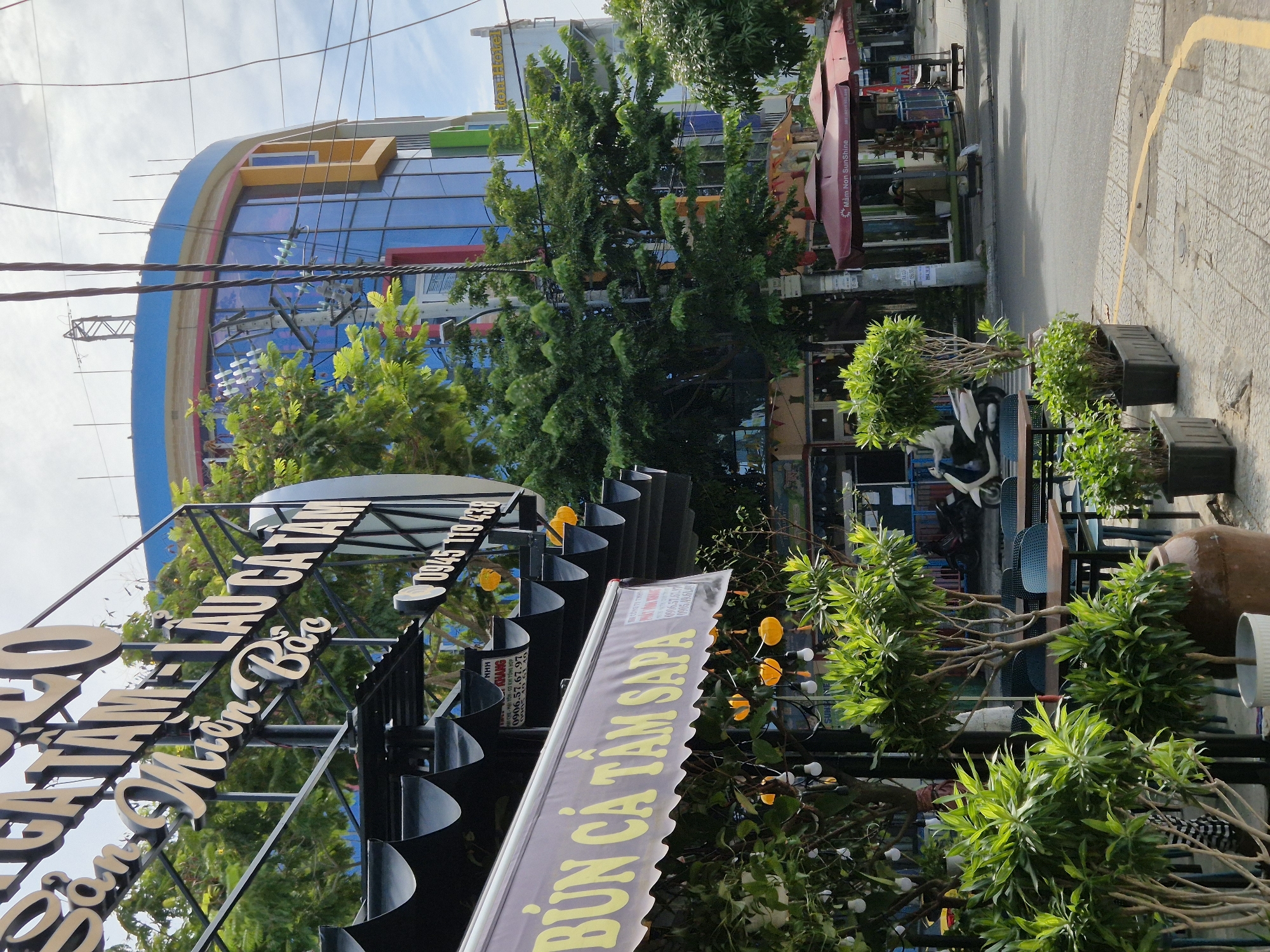 The image captures a street-level view of a commercial establishment in Da Nang, Vietnam. In the foreground, a restaurant called "Bún Cá Tầm Sapa" is visible, with its yellow lettering prominently displayed on a striped awning. To the left, potted green plants line the sidewalk, with outdoor seating tables and chairs set up behind them. A large, dark brown ceramic pot is also present.
Above the restaurant awning, a larger sign reads "Sàn Âm Thanh - Lẩu Cá Tầm - Miền Bắc." This sign, in white lettering, appears to be for a business that offers sound systems, fish hot pot, and northern Vietnamese cuisine. A phone number, "0945 119 438," is also visible on this sign.
In the background, a modern, multi-story building with a curved facade dominates the scene. The building features blue and yellow glass windows and appears to be brightly colored, with sections of red and green visible. A hotel sign, "Eden Hotel," is partially visible on the building.
The street is paved with concrete tiles and asphalt, with a yellow painted line marking the edge of the road. Several power lines and poles are present in the scene, crossing the sky. A motorcycle is parked on the right side of the street, and two red umbrellas with the text "Mầm Non Sunshine" are set up on the sidewalk further down the street, suggesting a nearby preschool or daycare. The sky is overcast, indicating a cloudy day. No people are clearly visible in the image, with the exception of a figure seated under one of the "Mầm Non Sunshine" umbrellas, whose identity is unclear.