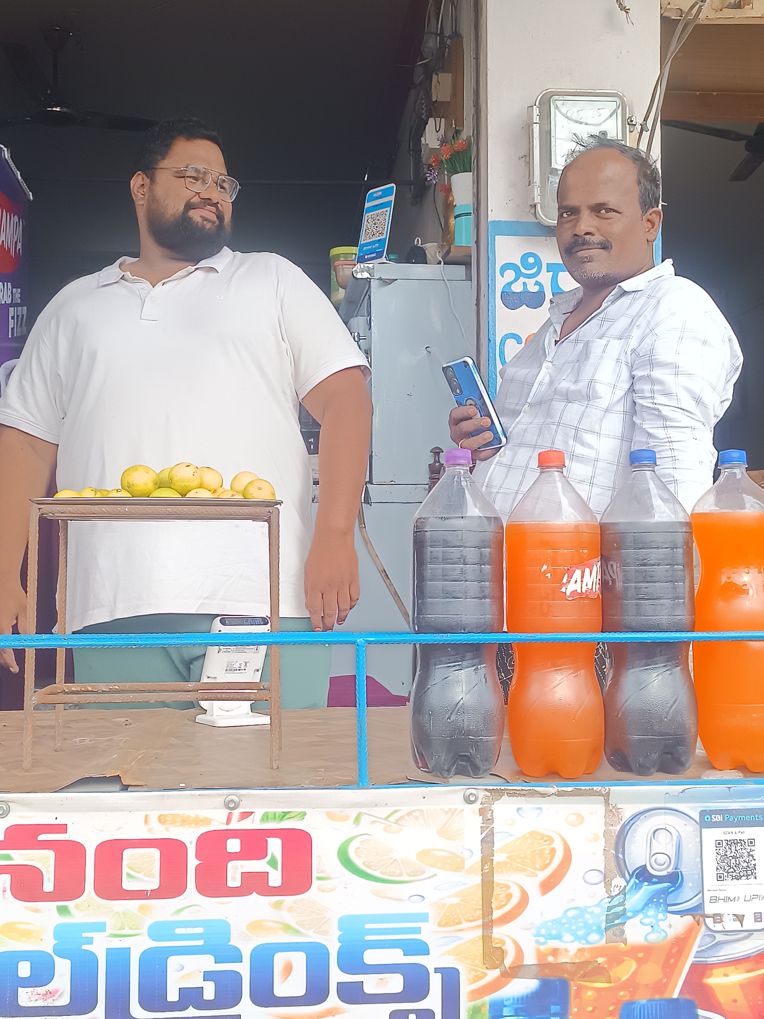 In Bethamcherla, India, two men are captured in a photograph at what appears to be a roadside stall. On the left, a man with a beard and glasses, wearing a white polo shirt, stands behind a display of lemons. A digital scale is visible beneath the lemons. To the right, a man in a checkered shirt holds a smartphone and stands in front of a selection of large soda bottles, some filled with a dark liquid and others with an orange beverage. Behind the men, a white electrical meter is mounted on a wall, and a colorful advertisement featuring fruit slices and beverage branding is prominently displayed at the bottom of the image. The scene suggests a commercial transaction or interaction at a local vendor.
