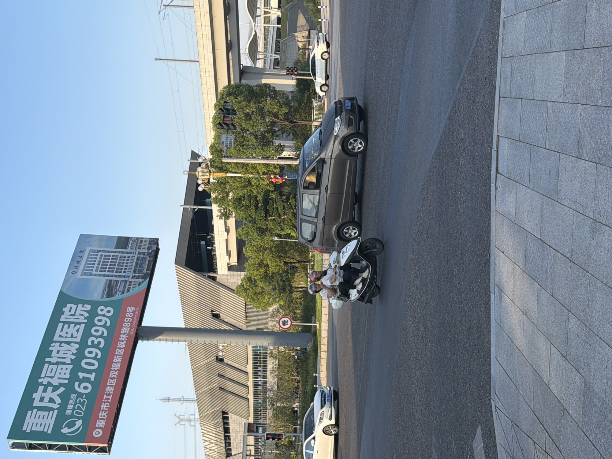 A wide shot captures a bright, sunny day at a busy intersection in Jiangjin, China, near Chongqing. Dominating the background is a large billboard for Chongqing Fucheng Hospital, featuring a phone number (023-61093998) and address in Jiangjin District. The billboard is mounted on a tall pole, with a modern architectural building visible behind it, possibly a transit station or commercial complex.
In the foreground, traffic moves through the intersection. A white sedan is visible on the left, and a dark gray minivan is in the center. A scooter carrying two passengers, both wearing helmets, is prominently featured in the middle right of the frame, moving through the intersection. The two passengers appear to be engaged in conversation or gesturing. Traffic lights are visible, indicating a regulated intersection. The road surface is asphalt, and a pedestrian walkway made of gray tiles is visible in the lower part of the image. Trees and overhead power lines are also present, adding to the urban landscape. The sky is a clear blue.