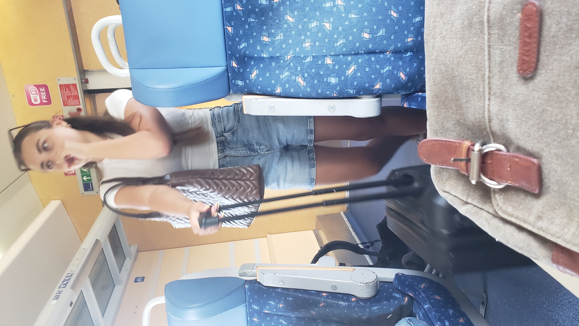 A young woman with dark hair, wearing a white t-shirt and denim shorts, stands on a train, holding a dark brown purse and a rolling suitcase. She is looking to the right and covering her mouth with her right hand. The train car has blue seats with a patterned fabric, and yellow walls. There are signs on the wall, including one that says "Wi-Fi FREE". The foreground shows a portion of a tan canvas bag with a leather strap and buckle.