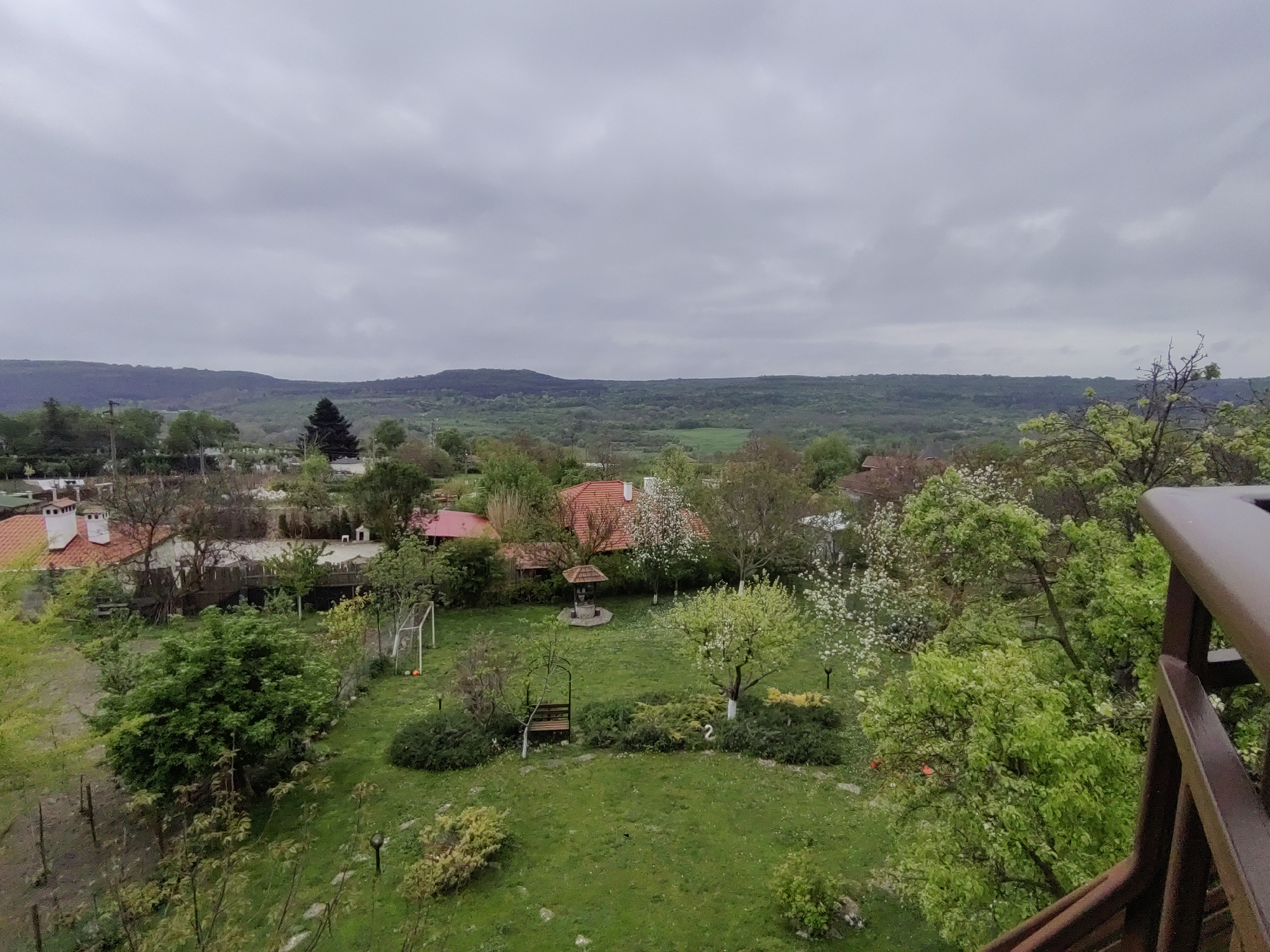 An eye-level, slightly aerial shot shows the backyard of a property in General Kantardjievo, Bulgaria, with a verdant lawn surrounded by trees, shrubs, and several buildings, under a cloudy sky. A wooden railing occupies the right side of the frame, slightly obscuring the view.
In the backyard, a grassy lawn dominates the scene. A seating area with a wooden bench is located in the center of the lawn, bordered by shrubbery. A covered well is visible. A soccer goal net stands on the lawn.
Red-roofed buildings are scattered in the background, interspersed with trees. Beyond the properties, forested hills extend horizontally across the horizon. The sky is overcast with dense clouds.