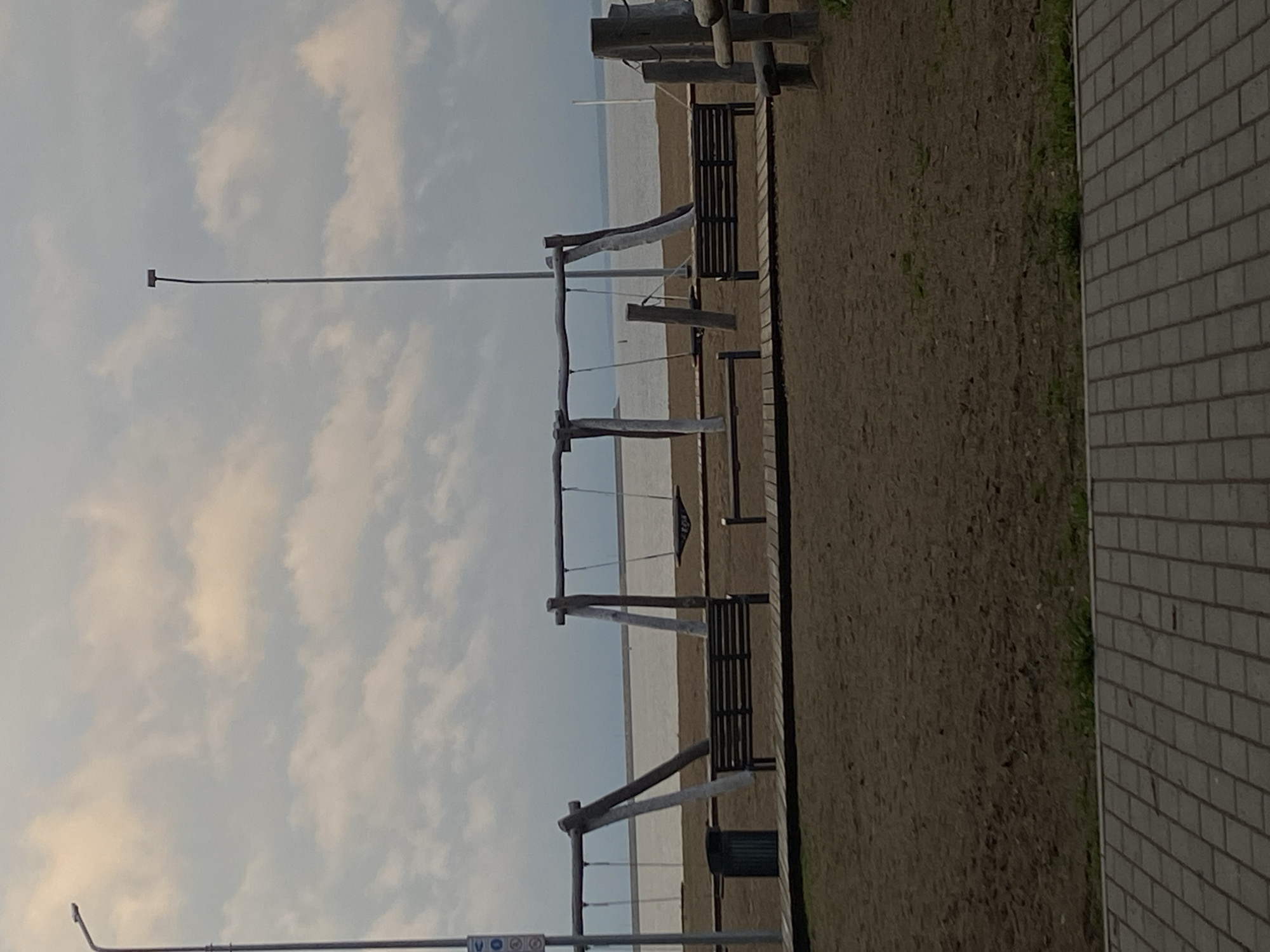 The image depicts a beach scene in what is presumed to be the city of Chaikovsky, Russia. A playground with swings is prominently featured, with several of the swings missing. The scene contains benches and a trash receptacle. Beyond the playground and beach, there is a body of water. The sky is overcast with white clouds. A brick wall borders the right side of the image. Two lampposts are also visible.