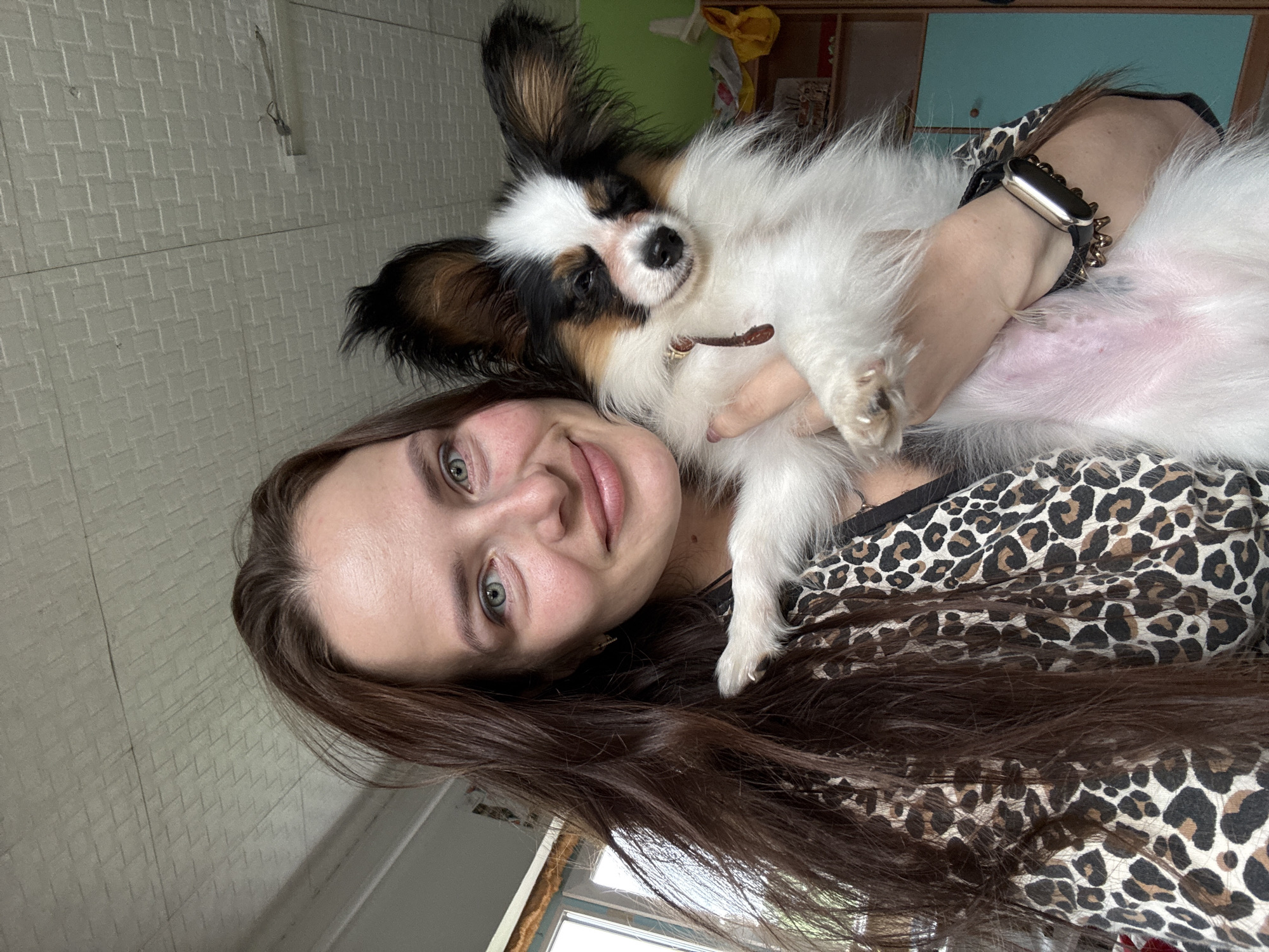 A young woman with long brown hair and green eyes is holding a small white and brown dog. She is wearing a leopard print top. The dog has large ears and is looking at the camera. The woman is smiling. The background shows a wall with a textured pattern and some furniture. The scene appears to be indoors, possibly a home, with natural light coming from a window. The overall mood is affectionate and casual, suggesting a moment captured between a pet and its owner. There is no visible text in the image. The photo was taken in Rusalevka, Russia.