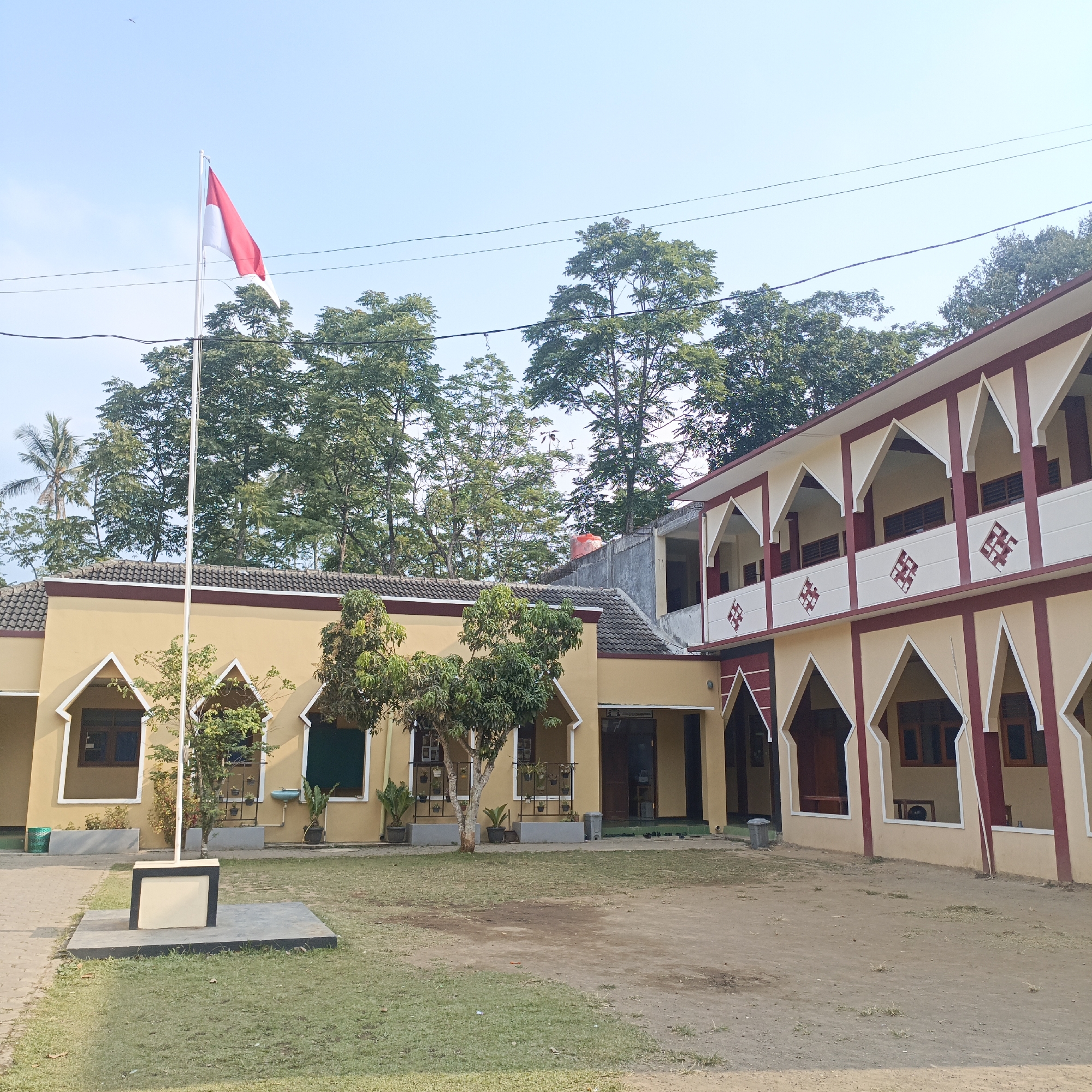 The image shows a schoolyard in Salatiga, Indonesia. In the foreground, a concrete flagpole base with a small rectangular plaque is visible on a patch of grass. The Indonesian flag, red and white, is flying from a tall flagpole to the left of the frame. Behind the flagpole, a light yellow building with a dark tiled roof extends across the middle ground. This building features arched windows and decorative trim. To the right, a two-story building with a similar architectural style, featuring arched openings on both levels and decorative diamond patterns, is present. The buildings are surrounded by greenery, with various trees visible in the background, including a palm tree to the far left. The sky is clear and light blue. The scene appears to be an outdoor educational institution.