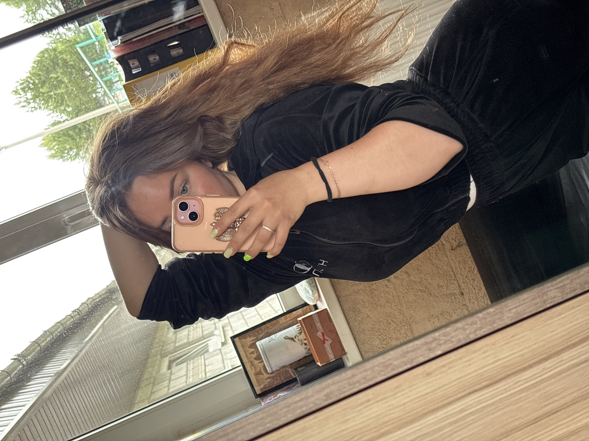 A young woman is taking a selfie in front of a mirror. She is wearing a black velvet tracksuit. Her hair is long and brown. She is holding a pink iPhone in her right hand, which has light green nail polish. She is wearing bracelets on her left wrist and a ring on her left ring finger. A photo frame and a box are reflected in the mirror. A window behind the woman shows trees and buildings. The city of Русский Вожой, Russia is not visibly indicated within the image.
