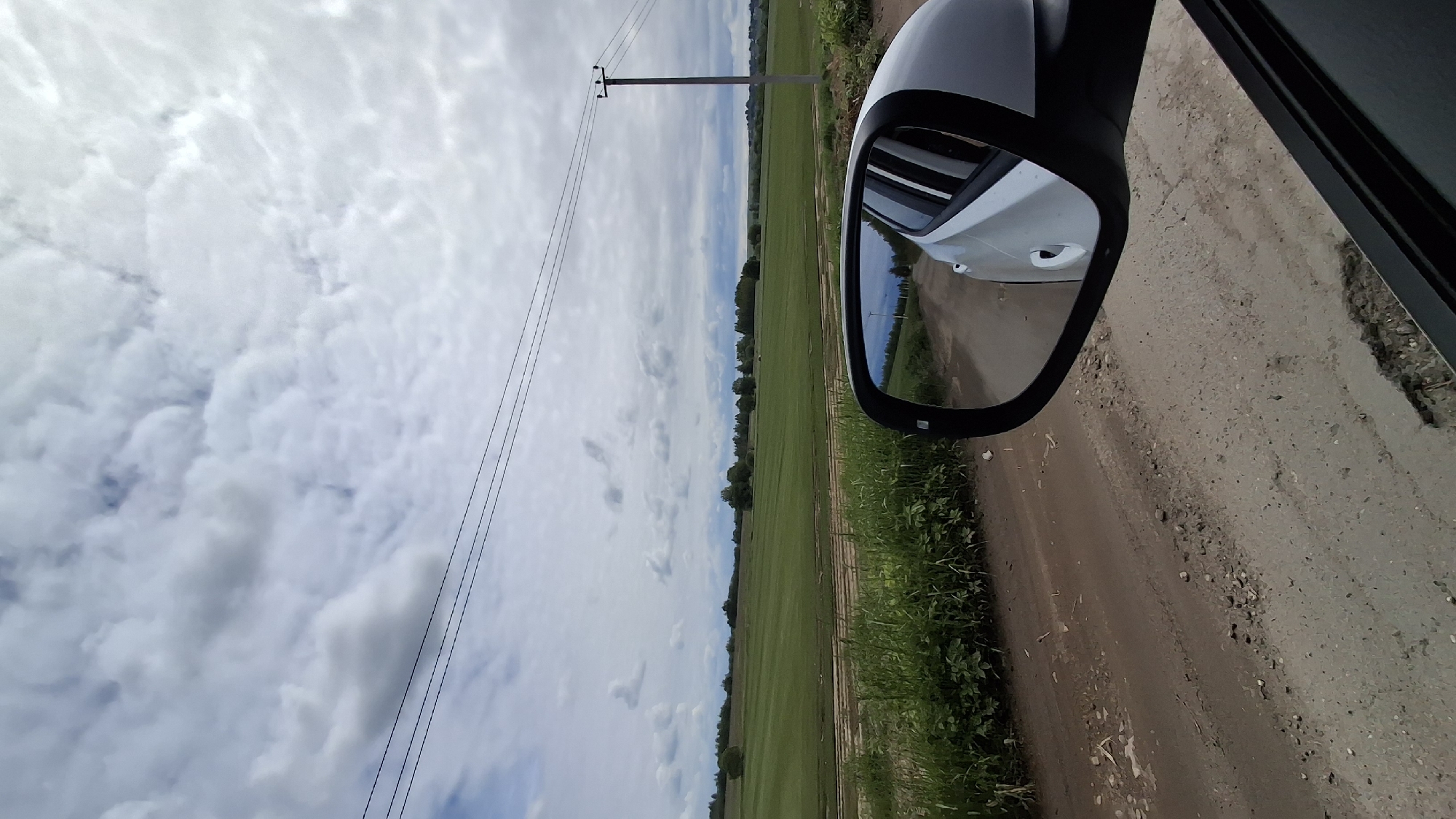 The image shows a view from a car driving on a dirt road in Innolovo, Russia. A side mirror of a white car is prominently featured. The landscape includes a field, trees, and a cloudy sky. Power lines are also visible. The road appears unpaved and somewhat uneven.