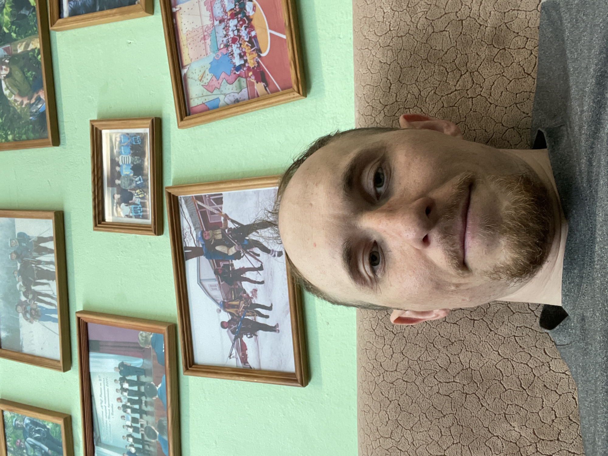 An adult male with a receding hairline, light eyes, and a light brown goatee is captured in an upward-angled selfie, looking directly at the camera with a neutral expression. He wears a dark grey t-shirt and is seated against a light brown, textured upholstered surface with a cracked pattern. The background consists of a uniformly painted light green wall. Nine wooden-framed photographs are affixed to the wall, depicting various group scenes: a group of approximately eight individuals in winter gear with poles in a snowy landscape; a single individual in camouflage clothing in a forest; a formal ceremony with individuals on a stage and an audience; a small group of five individuals in casual attire holding bags; a large group of children or young adults in a gymnasium with a colorful floor pattern; and another large photo showing a group of individuals in winter outdoor gear with backpacks and poles in a snowy, developed area with a building. The scene is indoors. Location: Unknown, Unknown.