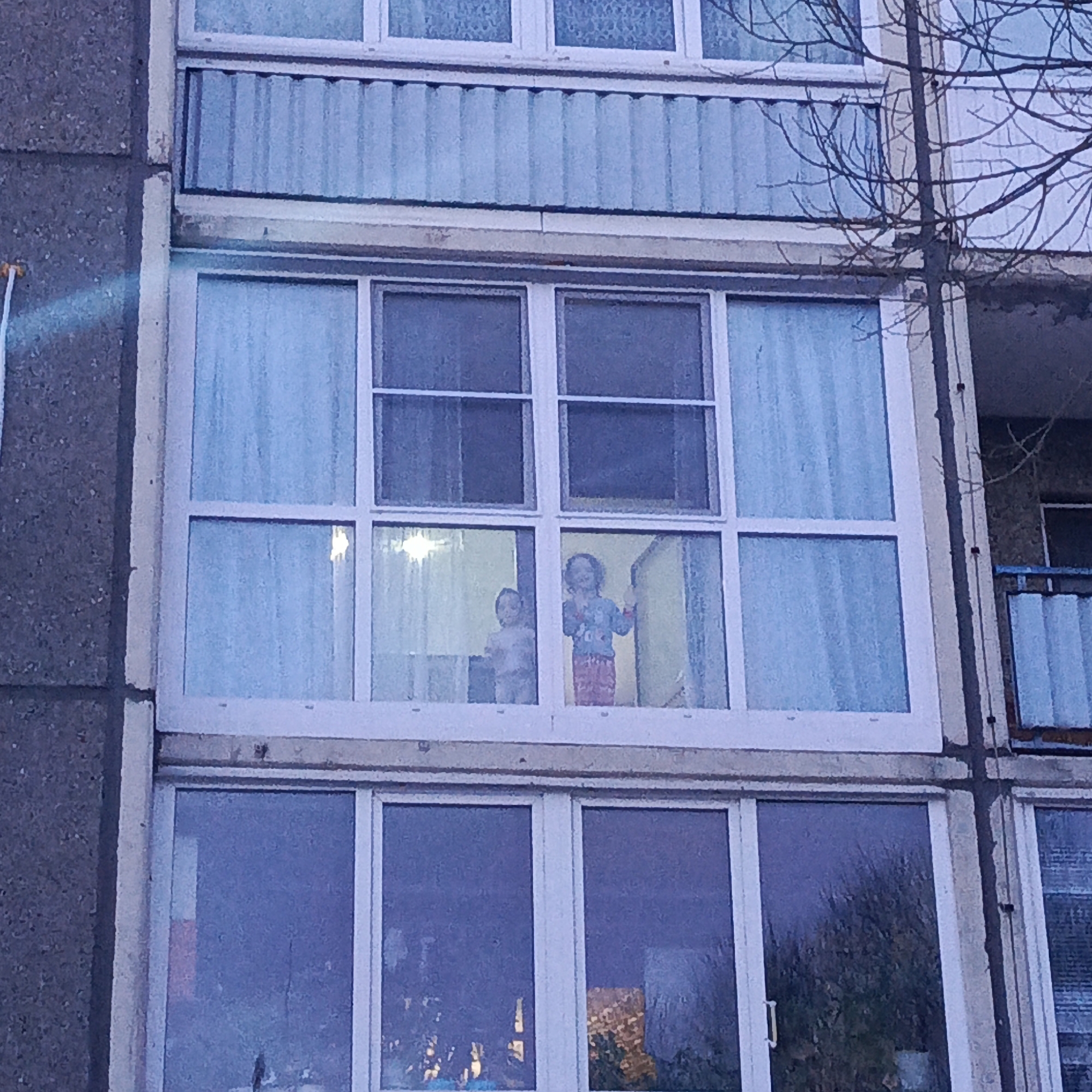 The image shows a section of a multi-story apartment building. The facade is primarily concrete, with white-framed windows. The middle floor window is the focal point, where two children are visible inside. One child is wearing red pajama pants with a blue shirt and is standing. The other child is wearing a white shirt and is also standing. The windows on the top and bottom floors have light blue curtains partially obscuring the view inside. A tree branch is visible on the right. Based on the query parameters, this scene is potentially located in Марковский, Russia.