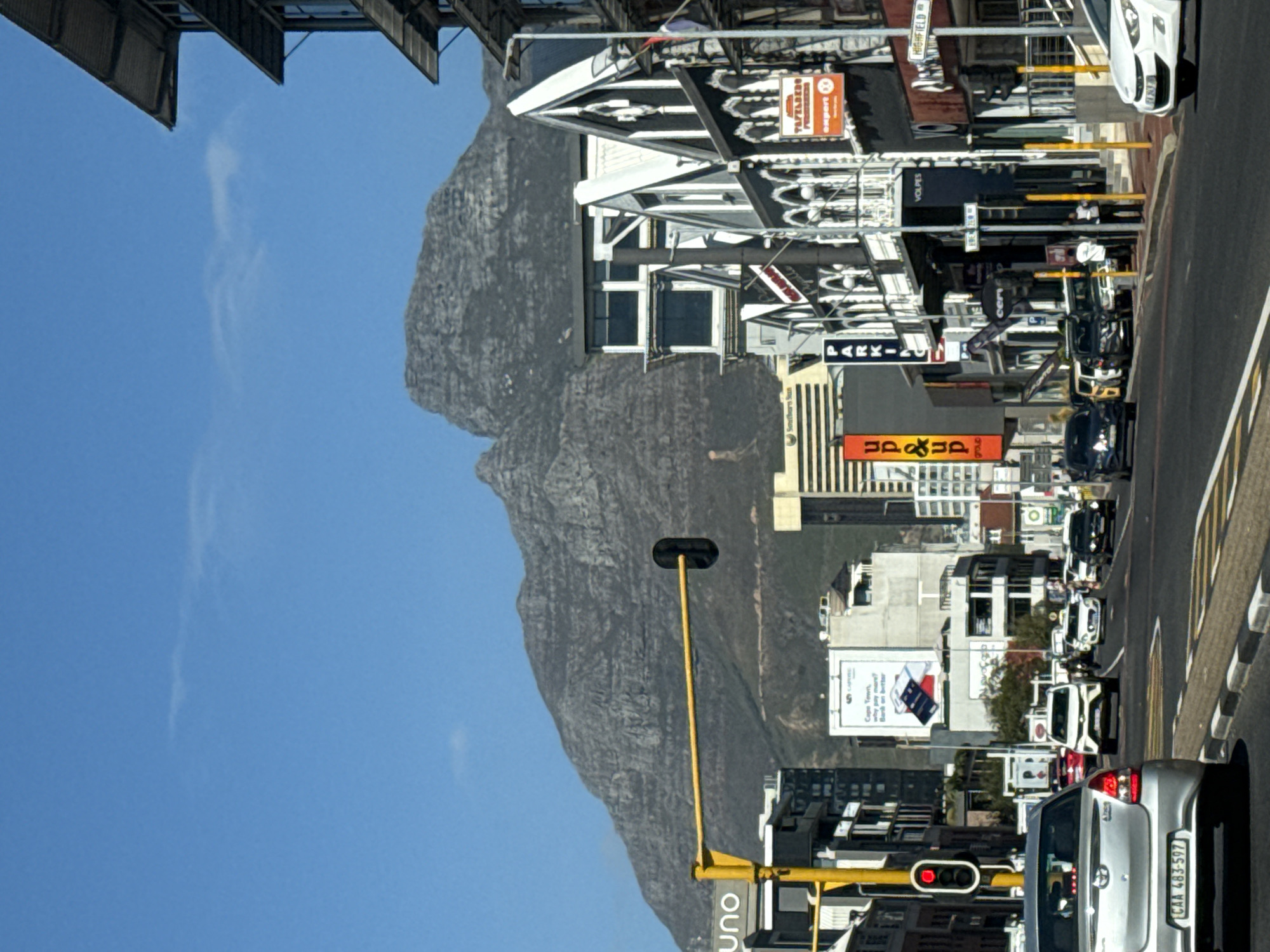 The image depicts a bustling city street scene in Cape Town, South Africa, with Table Mountain towering majestically in the background. The photograph is taken on a clear, sunny day, indicated by the bright blue sky and crisp lighting.
In the foreground, a silver car with the license plate "CAA 483-597" is prominently featured, stopped at a red traffic light. A yellow pole, possibly a streetlight or traffic signal pole, extends upwards. The street appears busy, with several other cars visible in the mid-ground, suggesting typical city traffic.
Along the street, buildings of varying architectural styles line both sides. Some structures feature ornate detailing, such as a black and white building with decorative white trim. Numerous commercial signs are visible, including "VOLPES," "HIGHFIELD," "PARKING," and "Up&up group." A partial sign reads "uno" and another "cerv." A billboard in the background advertises "Cape Town, why pay more? Bank on better," reinforcing the location. The "Southern Sun" logo is also partially visible on one of the buildings.
The dominant feature of the background is the rugged, grey rock face of Table Mountain, stretching across the horizon under a clear blue sky with only a faint wispy cloud. The overall scene captures the vibrant urban environment of Cape Town against its iconic natural landmark.