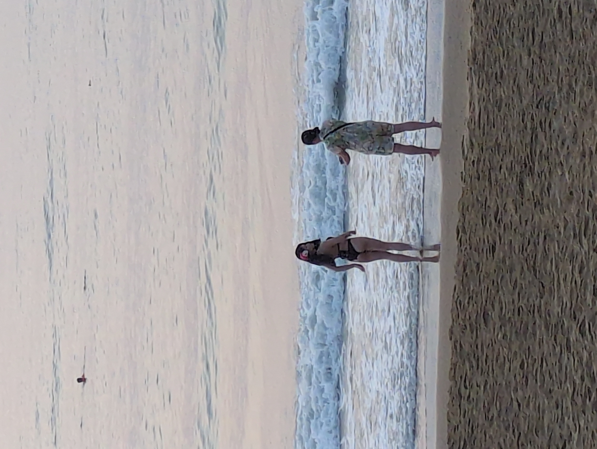 The image depicts a beach scene in South Quy Nhon Ward, Vietnam. A woman in a black bikini stands in the water near the shoreline, while a man wearing a patterned shirt and shorts stands further up the beach. The beach itself is a dark color. The ocean waves are visible, and the sky is overcast.