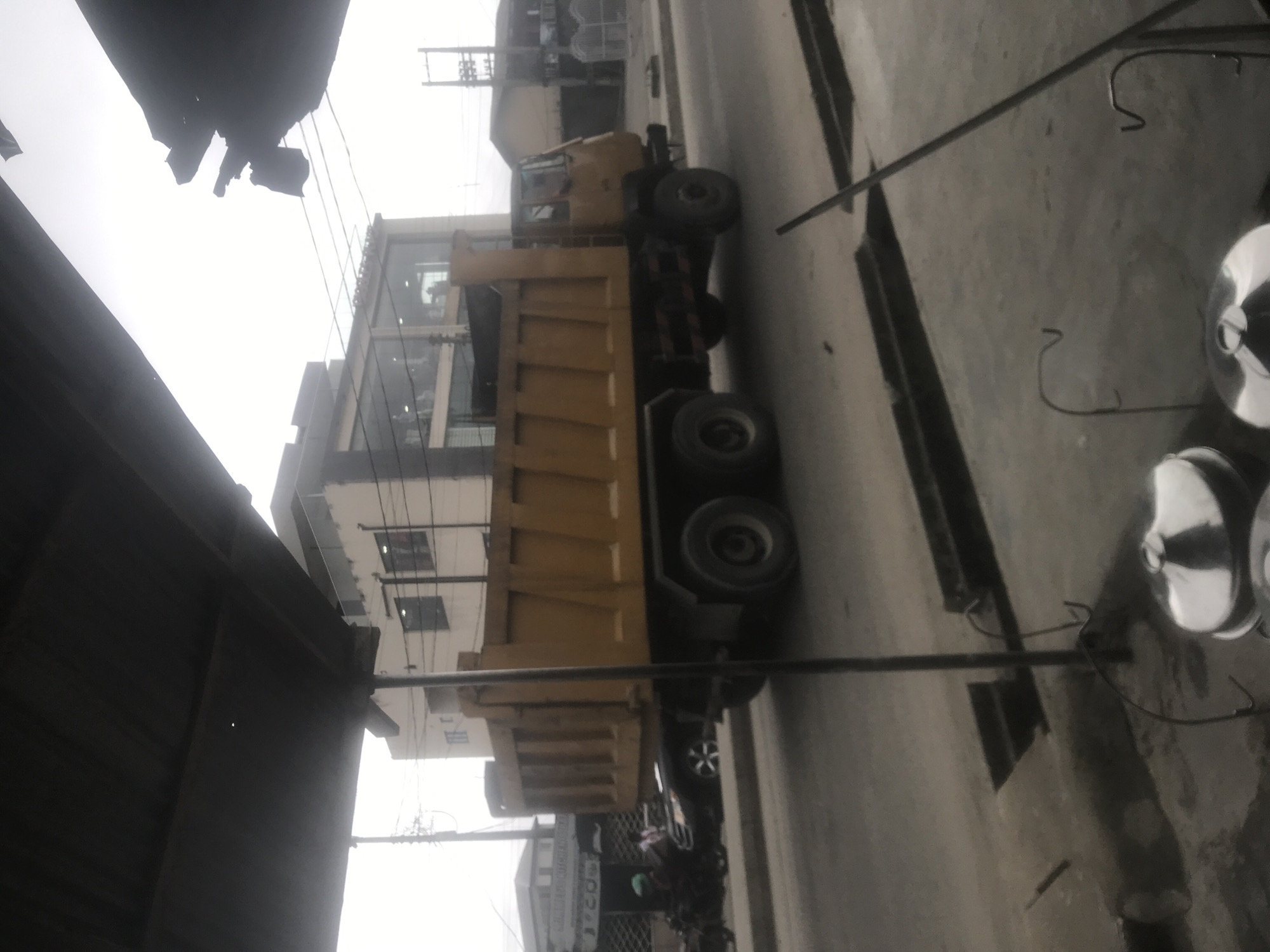 The image shows a yellow dump truck parked on a street. The truck is the main subject of the image, with its large yellow bed visible. The scene appears to be an urban environment, with buildings and power lines in the background. The sky is overcast, suggesting it might be a cloudy day.
To the left of the truck, a dark, slanted structure, possibly a roof or awning, partially obscures the view. In the background, a multi-story building with modern architecture and large windows is visible. The truck is positioned on a paved road, with a sidewalk or paved area to the right, where some metal objects, possibly cookware or industrial items, are placed.
The image is oriented with the truck running vertically, suggesting it was taken with the camera held sideways. The lighting is diffused, consistent with an overcast sky, and there are no strong shadows. There is no visible text in the image. The overall impression is one of a street scene with a parked truck, possibly in a commercial or industrial area. The weather appears to be dull or cloudy.