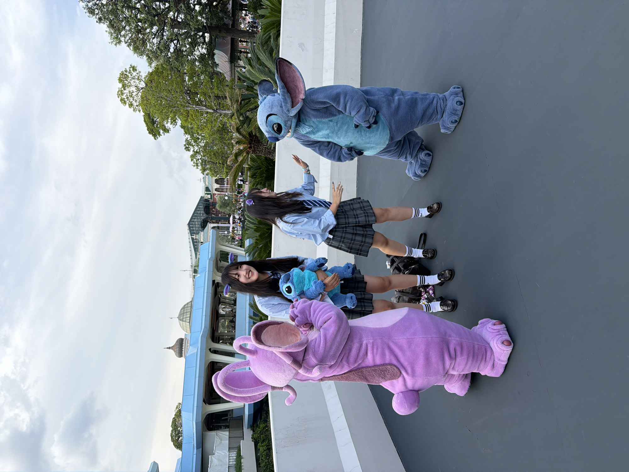 In Urayasu, Japan, two young women in school uniforms interact with costumed characters from Disney's Lilo & Stitch. On the left, a person in a large purple Stitch-like costume stands with its back to the camera. To its right, a young woman holding a blue Stitch plush toy smiles at the purple costume. Between the two young women, a second young woman in a similar school uniform interacts with a person in a blue Stitch costume, extending her hand towards it. Both women are wearing plaid skirts, white shirts, and knee-high socks with loafers. The Stitch costume appears to be interacting with the woman by raising its hand. The scene takes place outdoors on a dark gray paved surface with a white railing in the background. Beyond the railing, buildings with futuristic architecture, trees, and a cloudy sky are visible. The overall atmosphere suggests a theme park environment.