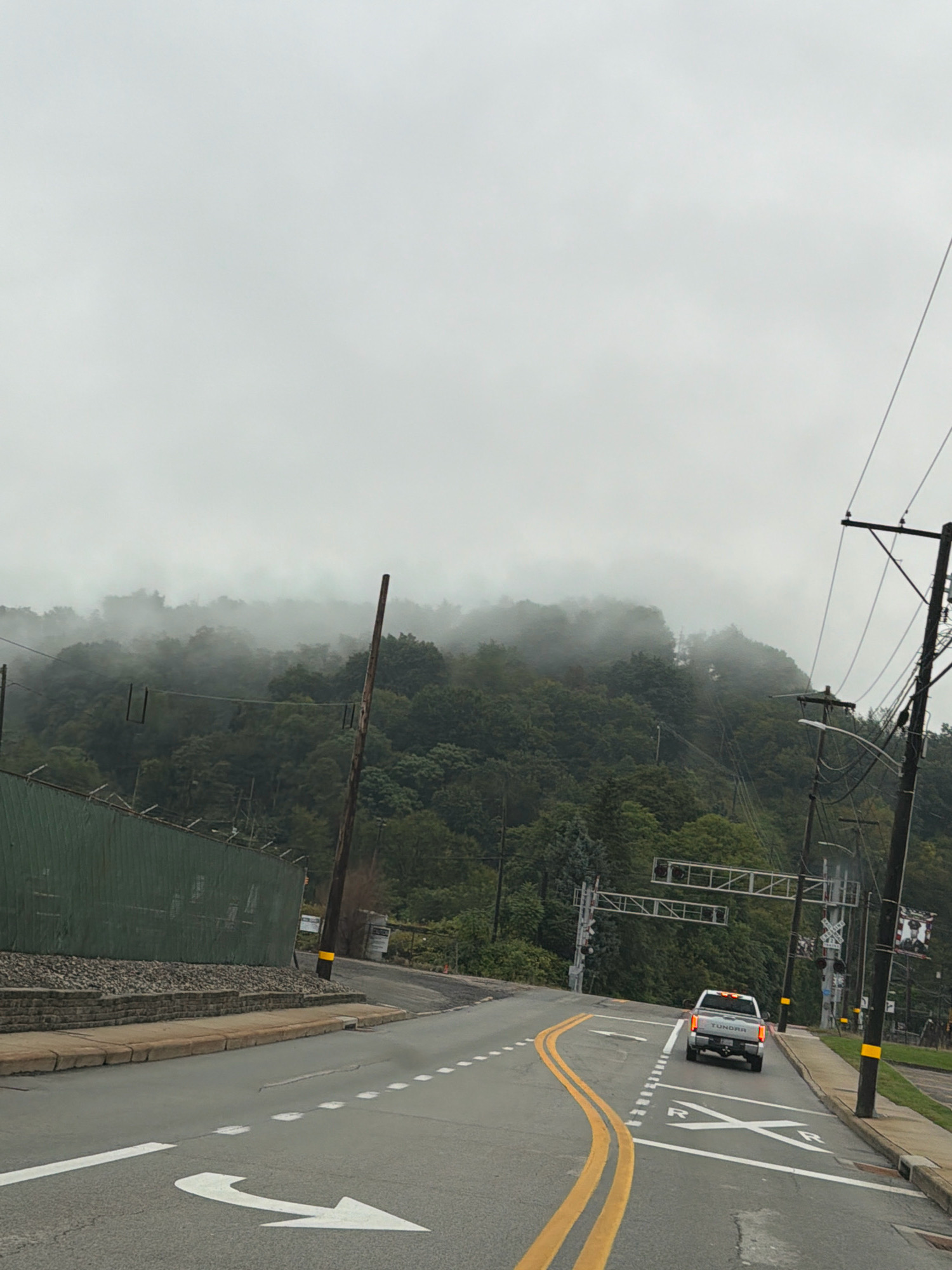 The image depicts a road scene in Weirton, United States of America, on an overcast day with fog present. A silver pickup truck, identified as a TUNDRA, is driving on the road, which has yellow double lines and white lane markings including a right-turn arrow and a crosswalk marking with "RR" and "X". To the left of the road, there is a green fence with a barbed wire top, followed by a retaining wall and a sidewalk. A wooden utility pole with attached wires and a yellow band is visible on the right side of the road. Further in the background, there are more utility poles and wires, and a forested hillside partially obscured by fog. A set of railroad crossing signals are also visible.