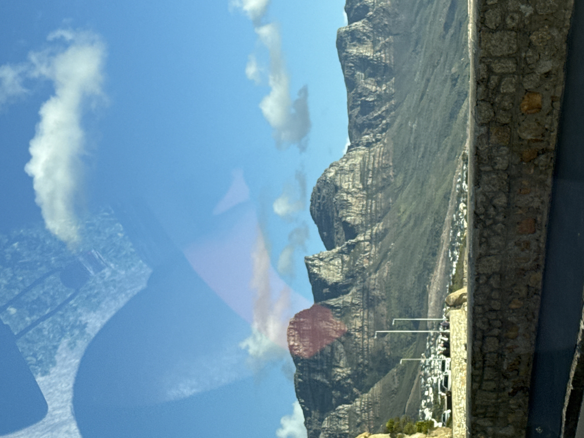 The image shows a view of Table Mountain in Cape Town, South Africa, partially obscured by a reflection. The sky is blue with scattered white clouds. A rocky, green-covered mountain dominates the view. A portion of the mountain is obstructed by an internal reflection of car equipment. A stone wall runs along the right edge of the image, partially obscuring a view of what appears to be residential area. The reflection makes it difficult to completely discern the environment.
