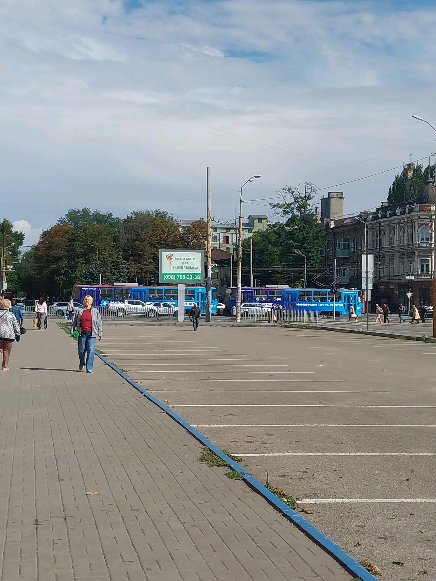 The image depicts a sunny day in a city, likely Dnipro, Ukraine, given the Cyrillic text on the advertisement. A paved pedestrian walkway runs along the left side of the frame, where several individuals are walking. A woman in a red top and blue jeans is walking towards the viewer, carrying a green bag. Behind her, other pedestrians are also walking along the sidewalk.
In the background, a wide street or square is visible with a parking lot marked by white lines. Several blue trams are present on the street. One tram in the mid-ground displays an advertisement with Cyrillic text and a phone number, "(098) 788-33-38". The advertisement also features an image of a rose.
Further in the background, buildings of various architectural styles are visible, including some older, ornate structures with intricate details, and more modern buildings. Trees with green and autumnal foliage are scattered throughout the scene. The sky is partly cloudy with a mix of blue sky and white clouds. Streetlights are positioned along the walkway and street. The overall scene suggests a public urban space with ongoing pedestrian and public transportation activity.