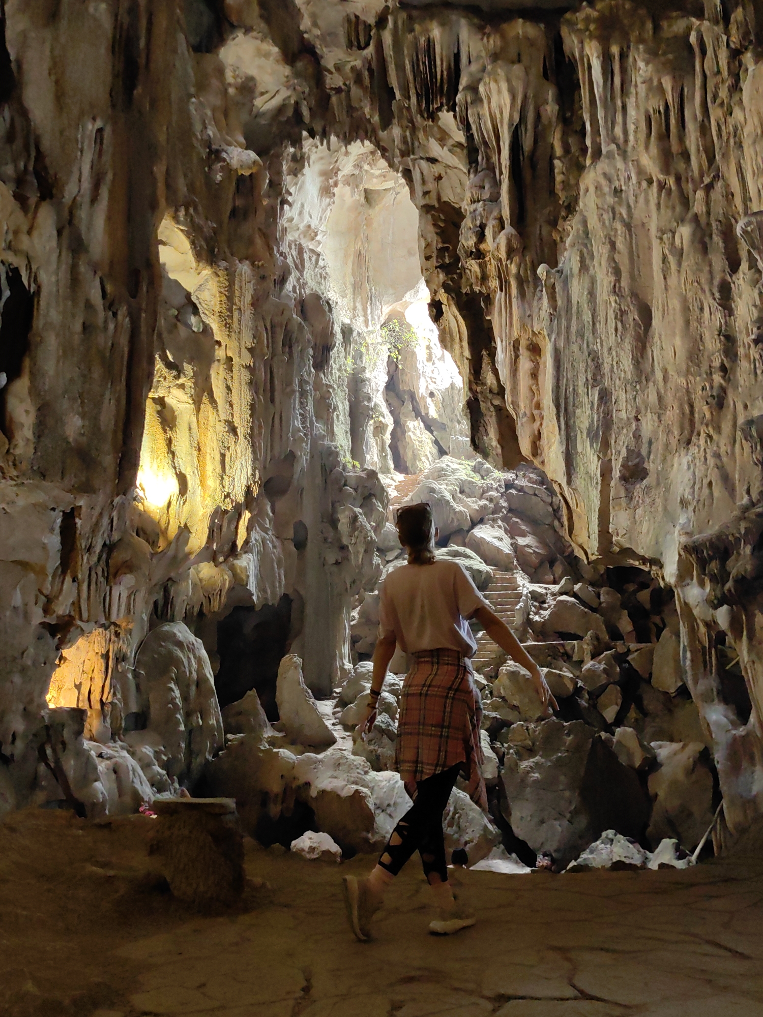 A wide-angle shot depicts a person in the Tam Thanh Cave in Lạng Sơn, Vietnam. The person, seen from behind, wears a light-colored t-shirt, a plaid shirt tied around their waist, black leggings with cutouts, and light-colored shoes. They stand with arms outstretched, seemingly balancing as they walk towards a steep, rocky path that leads upwards.
The cave environment is characterized by stalactites and stalagmites of various sizes, with some illuminated by artificial lighting to create a warm, yellow glow. Natural light streams through an opening at the top of the cave, highlighting the rocky terrain. The ground near the viewer is relatively flat, transitioning to a more uneven, rocky surface further into the cave.