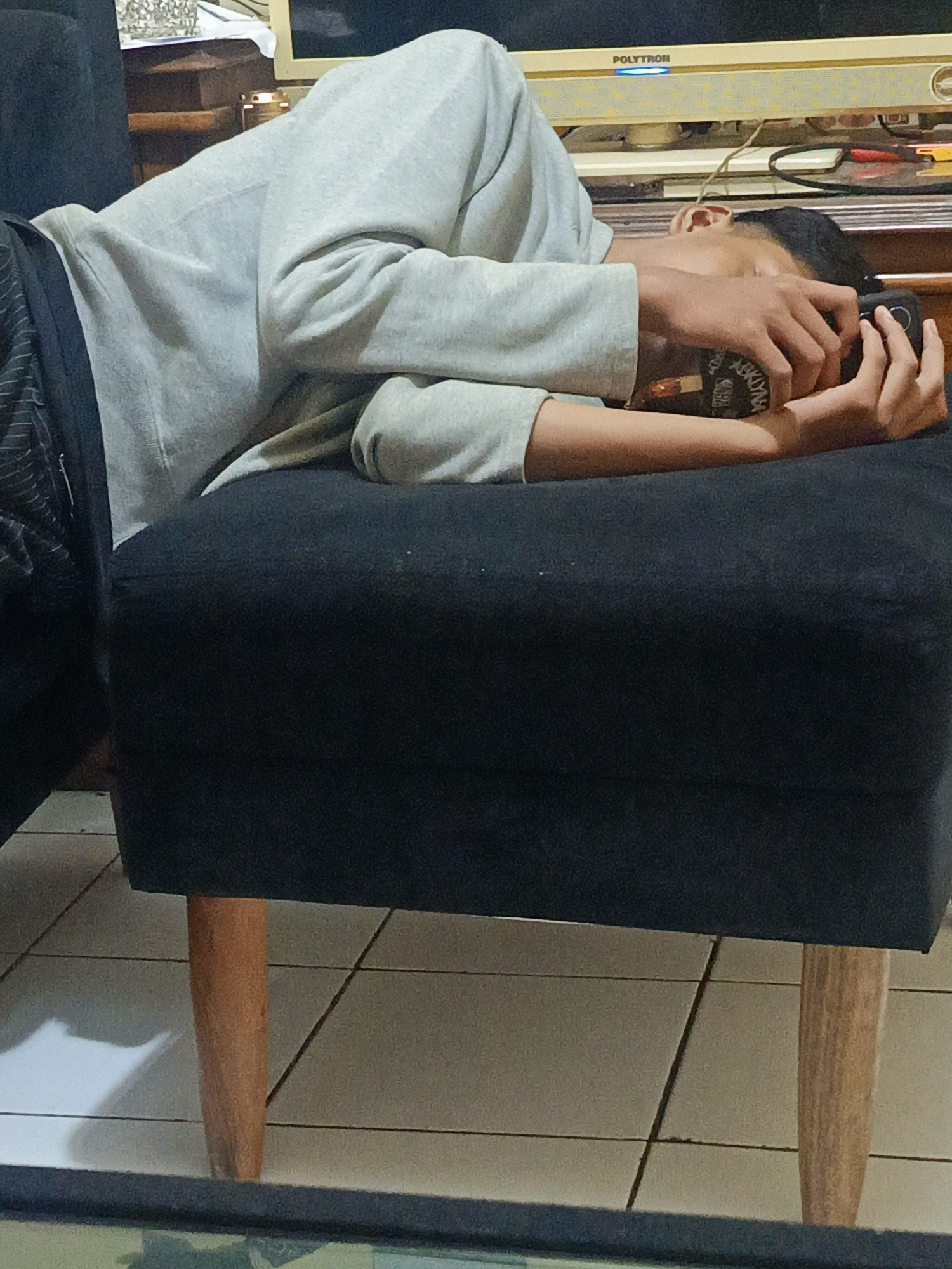 A young male is lying on a dark ottoman with wooden legs. He is wearing a light gray long-sleeved shirt and dark pants with vertical stripes. He is holding a dark-colored object, possibly a phone or a camera, up to his face and appears to be taking a picture or recording. His face is partially obscured by his hand and the object. The ottoman is positioned on a tiled floor. In the background, a television screen with the word "POLYTRON" is visible. The overall setting suggests an indoor environment. The image was taken in the city of Margajaya, Indonesia.