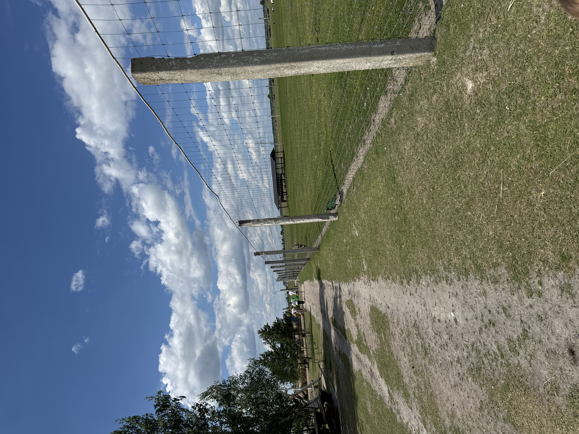 The image depicts a rural outdoor scene under a blue sky with white clouds. In the foreground, a dirt path runs alongside a wire fence supported by concrete posts. The path is bordered by grass on both sides. Several trees are visible to the left of the path, with some picnic tables and hay bales in the background. Further down the path, a group of people are walking away from the camera. Beyond the fence, a large green field stretches out, containing a covered structure with several animals nearby, possibly cattle. The overall setting appears to be a farm or a rural park. The location is identified as Kostiantynivka, Ukraine.