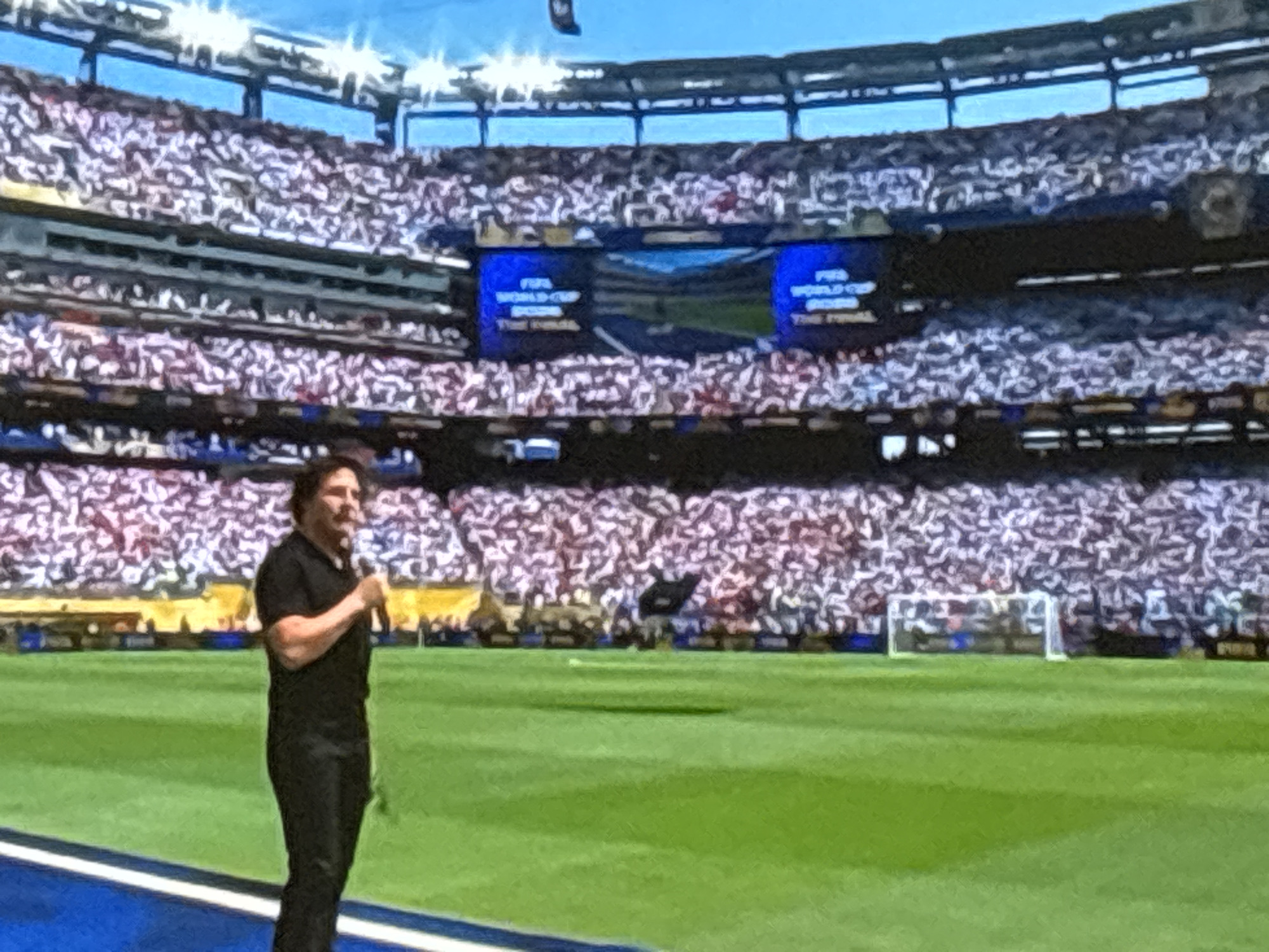 The image shows a man speaking into a microphone on a soccer field. He is wearing a black shirt and pants. Behind him, a stadium is filled with spectators. Large screens display "FIFA WORLD CUP". The scene is set in a large stadium, and the presence of FIFA branding suggests a sporting event. The man appears to be addressing the crowd, possibly as a presenter or performer. The stadium's architecture indicates it is a modern sports venue. The image does not contain any visual cues that would allow for the identification of the city as Burgas, Bulgaria.