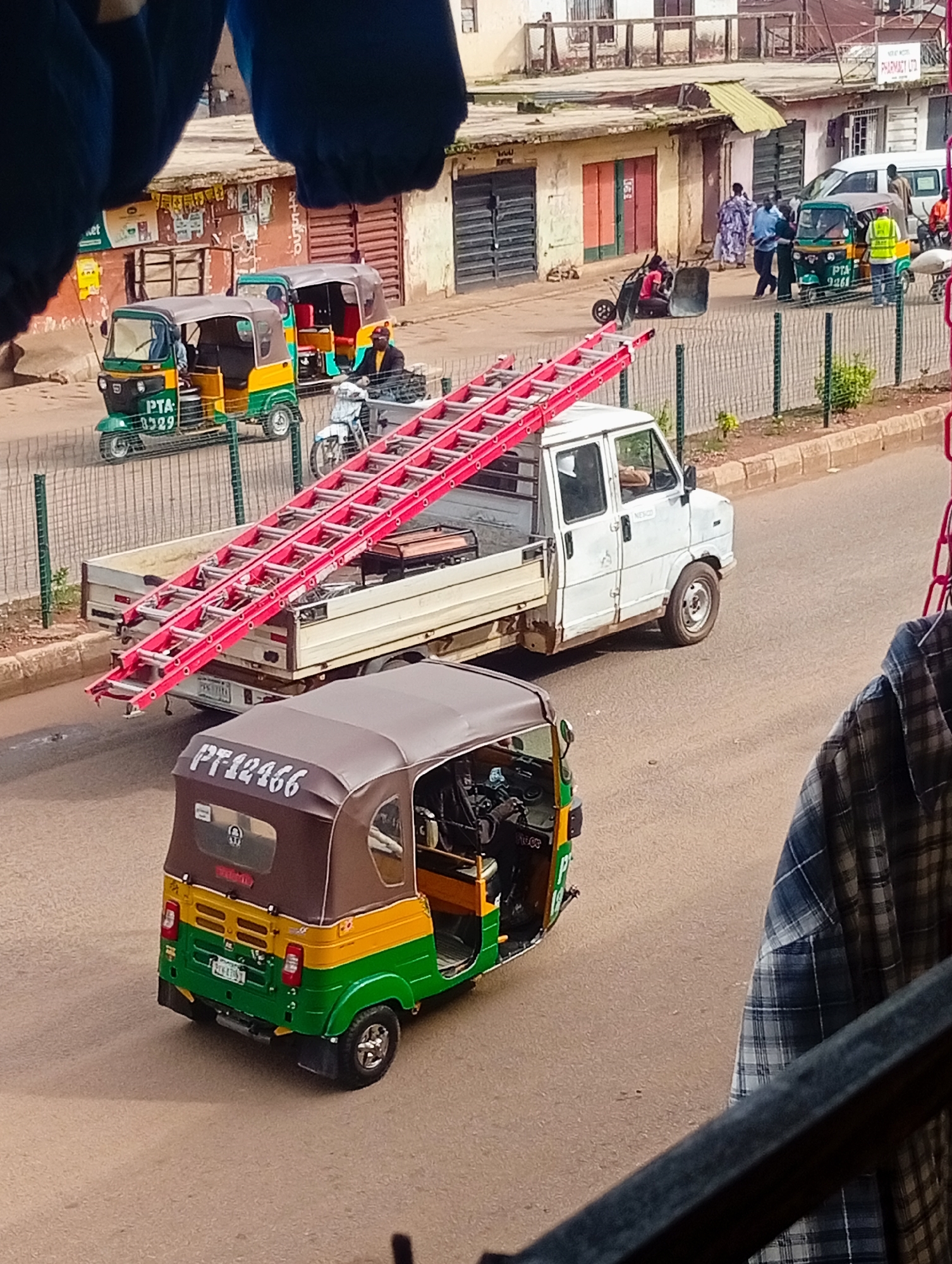 The image depicts a street scene in Bukuru, Nigeria. In the foreground, a brown and green motorized tricycle, identified by the license plate "PT12166," is parked or moving on the asphalt road. To the right, a portion of a blue and white plaid shirt is visible, hanging perhaps from a railing or balcony.
In the mid-ground, a white flatbed truck is carrying a long, bright red ladder. Behind the truck, a green and yellow motorized tricycle with the license plate "PTA 9229" is visible. Further back, there are other tricycles and a white van. People are seen near the buildings, some appearing to be pedestrians and others possibly drivers or vendors. The buildings in the background are typical of a commercial area, with storefronts and signs, some with signs of age and wear. The overall environment suggests a busy urban setting with vehicular and pedestrian activity. A metal fence runs along the edge of the road.