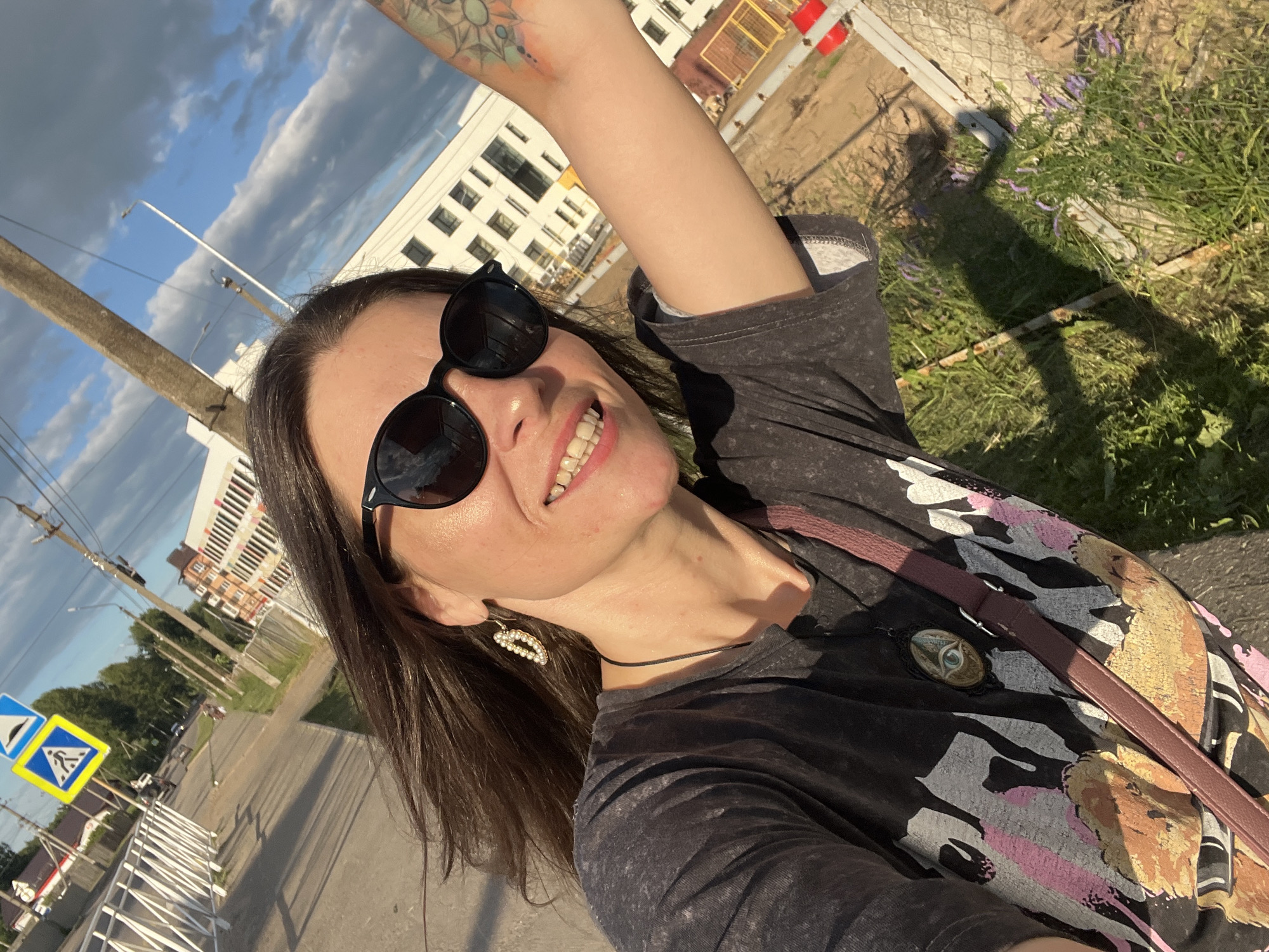A woman is taking a selfie outdoors, smiling at the camera. She is wearing sunglasses, a dark t-shirt with a graphic print, and a thin black choker necklace. She also has a bag with a burgundy strap slung across her body. A notable detail is a tattoo on her raised arm. The background shows a street scene with a modern building, a lamppost, road signs, and vegetation. The sky is cloudy with patches of blue, indicating it could be daytime with variable weather. The overall mood appears cheerful and relaxed. There is no visible text in the image.