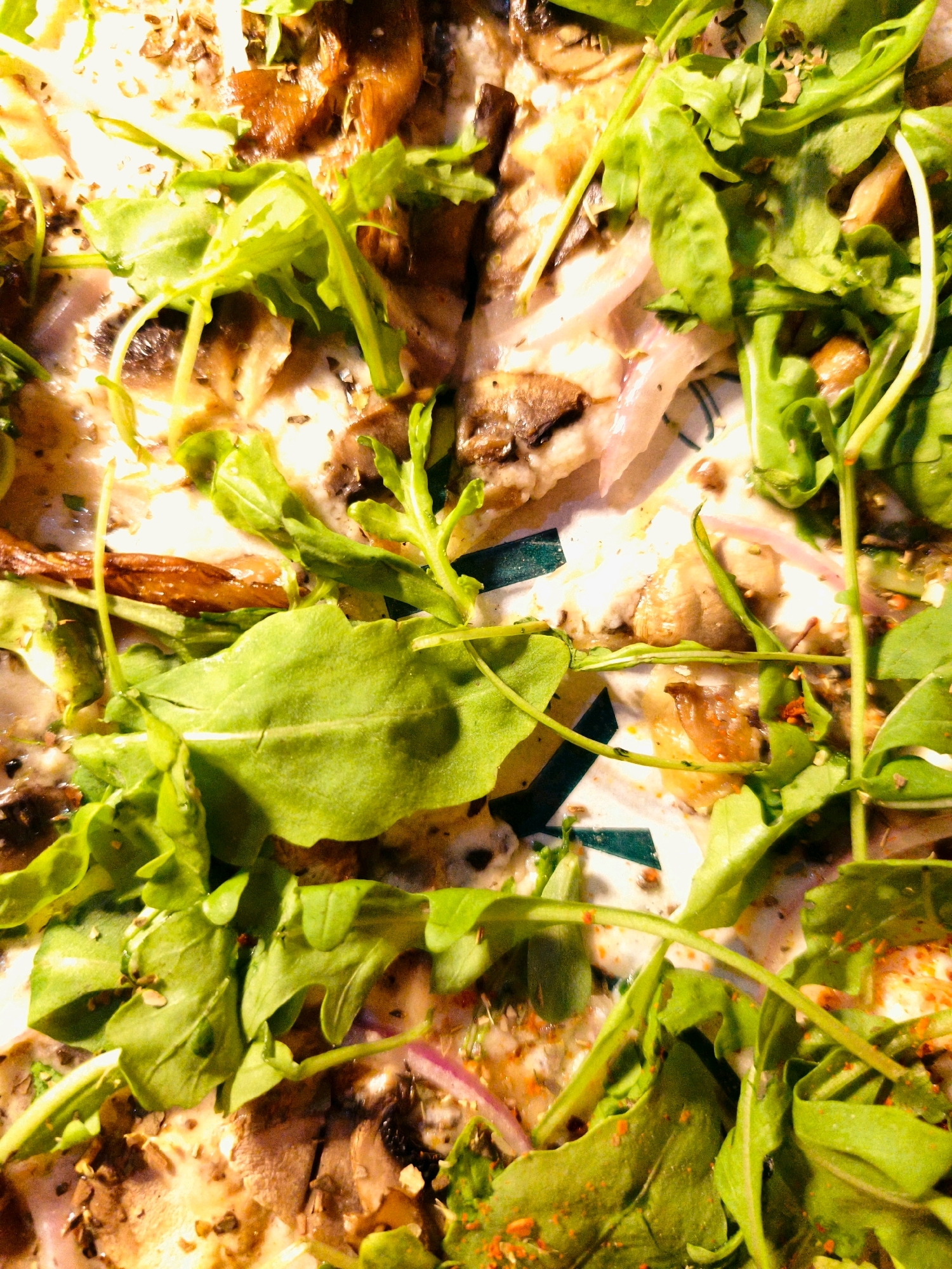 A close-up, high-angle shot reveals a visually rich pizza, heavily garnished with a generous amount of fresh arugula. The arugula's vibrant green leaves, some curled and delicate, others broad and flat, are scattered unevenly across the surface. Interspersed with the greens are thinly sliced mushrooms, their earthy brown tones contrasting with the lighter ingredients. Hints of red onion, appearing as translucent slivers, add another layer of color and texture. The pizza base is visible in places, covered in melted cheese, and a scattering of dried herbs and red pepper flakes adds further visual complexity and a sense of spice. The lighting casts a warm glow on the food, highlighting the textures and colors of the ingredients. No people or specific location within Be'er Sheva, Israel are identifiable.