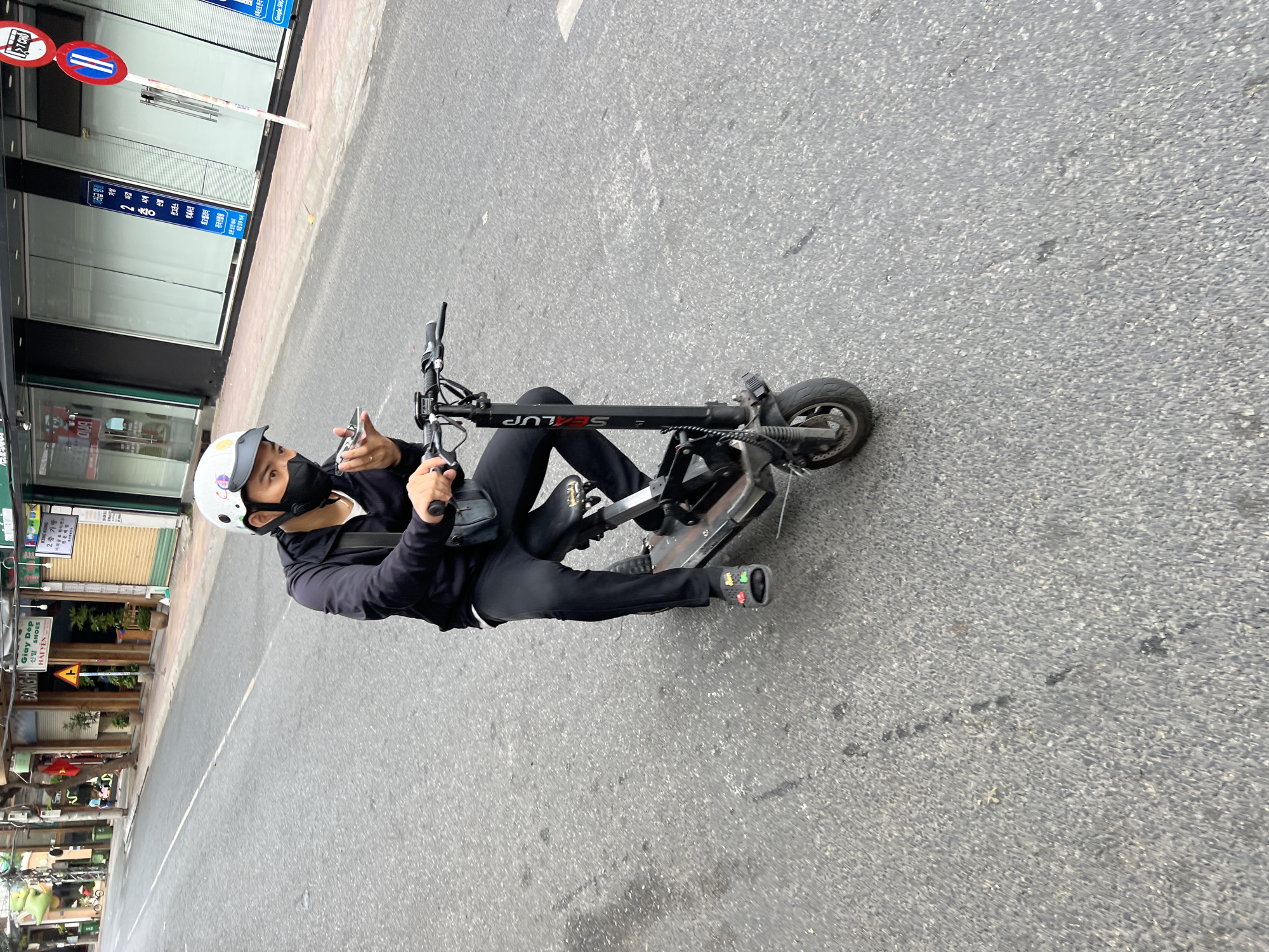 An Asian male wearing a black face mask, a black zip-up hoodie, black pants, and a white helmet with blue and grey decals is riding a black electric scooter on a paved road in Nha Trang, Vietnam. He is holding a silver smartphone in his right hand, appearing to be speaking into it. The scooter has the brand name "SEALUP" printed vertically on its stem. He is wearing colorful slide sandals. The road appears to be a city street with buildings lining the right side. The buildings have storefronts with signage in Vietnamese and possibly Korean. There are traffic signs visible, including a red and white circular sign indicating no entry, and a yellow triangular sign with a black arrow. The weather appears overcast.