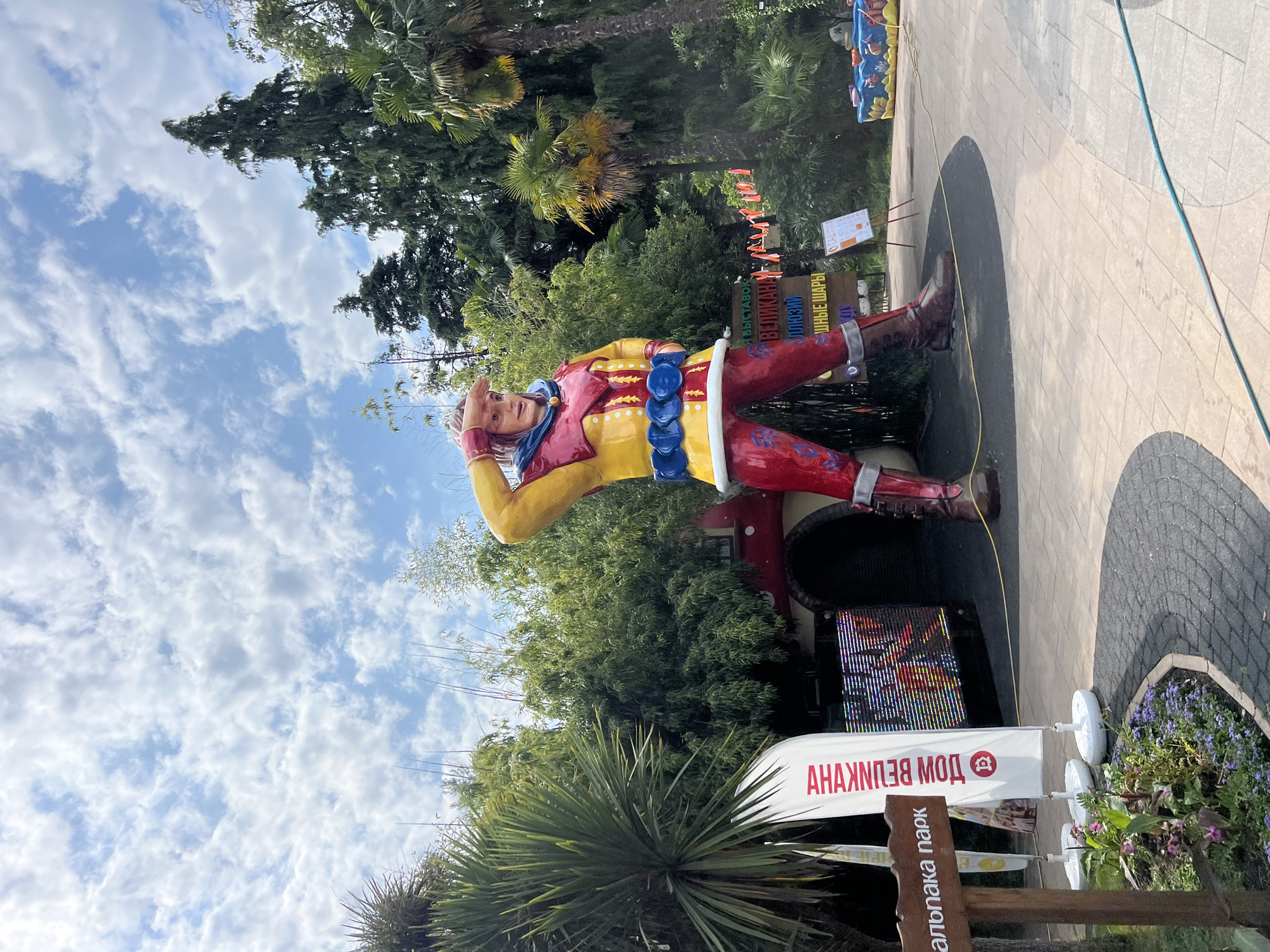 The image shows a large statue of a man dressed in a yellow and red costume, standing in an outdoor amusement park. The statue is in a heroic pose, with one hand shielding its eyes as if looking into the distance. The statue is located in the city of Sochi, Russia, and is situated in front of a building with a red facade and an arched entrance.
Surrounding the statue are lush green trees and palm trees, suggesting a warm climate. Several signs are visible in the foreground and background, some with Cyrillic text. One sign, to the left of the statue, appears to be a wooden directional sign with the words "МАЛПАКА ПАРК" (Malpaka Park). Another sign, a white banner to the right of the wooden sign, has red text that reads "АНАНУШКА" (Ananushka) and a circular emblem. Other signs further in the background contain text that appears to be advertising attractions.
In the background, there's a large LED screen displaying colorful graphics, and a small, brightly colored ride or attraction with a blue and yellow base. The ground is paved with light-colored interlocking stones, with a darker curved border around a flowerbed in the lower left corner. The sky is blue with fluffy white clouds. The overall scene depicts a vibrant and family-friendly amusement park environment.