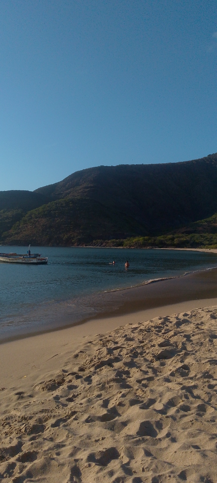 A coastal scene depicts a serene beach with golden sand marked by numerous footprints. The calm, clear water extends towards a backdrop of lush green hills under a clear blue sky. A small boat is anchored near the shore, and two individuals are seen swimming in the water, creating a peaceful, summery atmosphere. The scene evokes a sense of relaxation and tranquility.