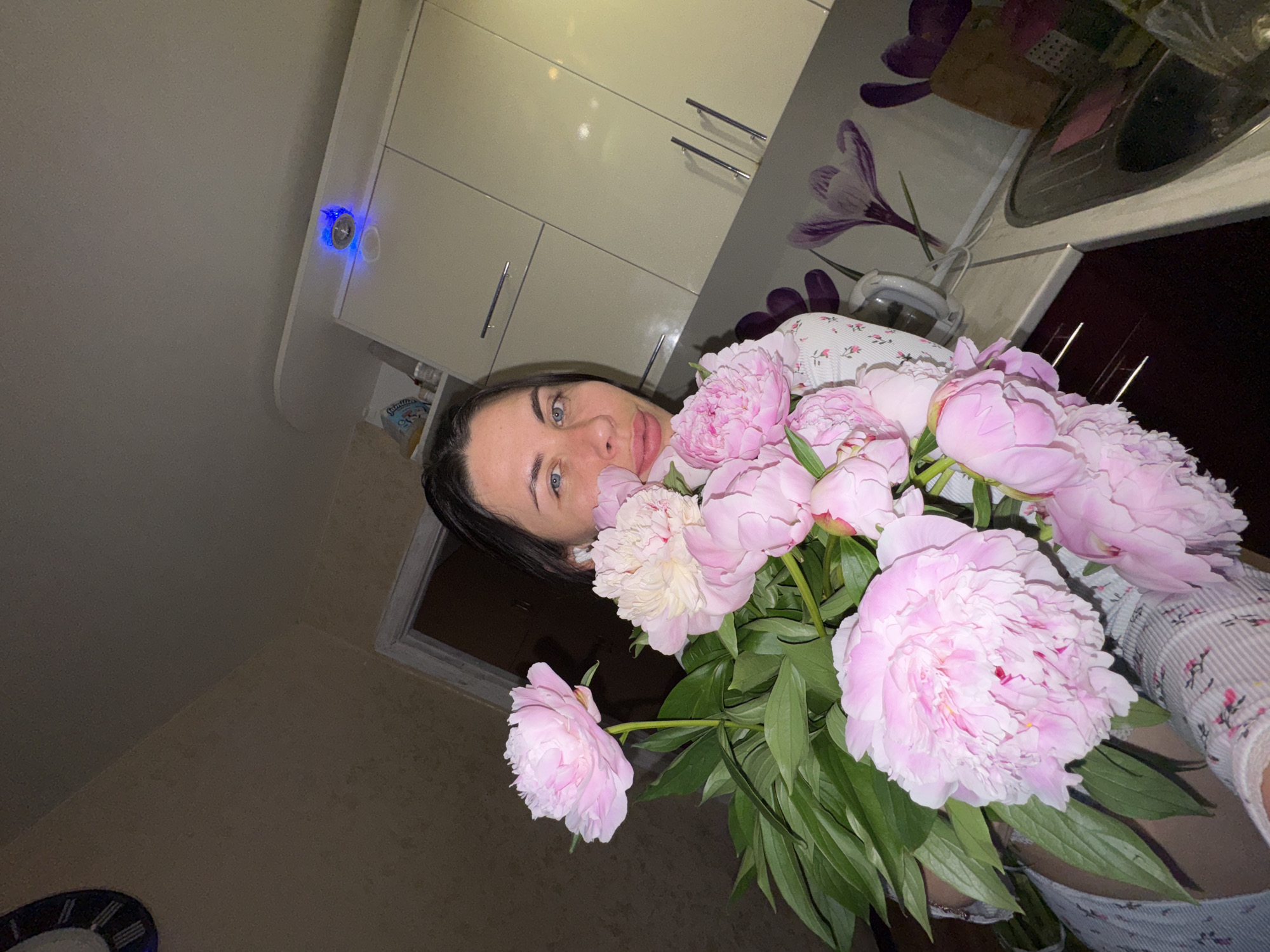 In a kitchen, a woman of European descent with dark hair and blue eyes is holding a bouquet of pink peonies. She is wearing a light-colored shirt with a floral pattern. The kitchen features light-colored cabinets, a countertop with a sink, and a backsplash with a floral design. A blue LED light is visible on top of the cabinets. The overall lighting suggests it is nighttime or indoors. There is a clock on the wall to the left. The image does not provide any specific identifying details about the location within the city of Chaikovsky, Russia.