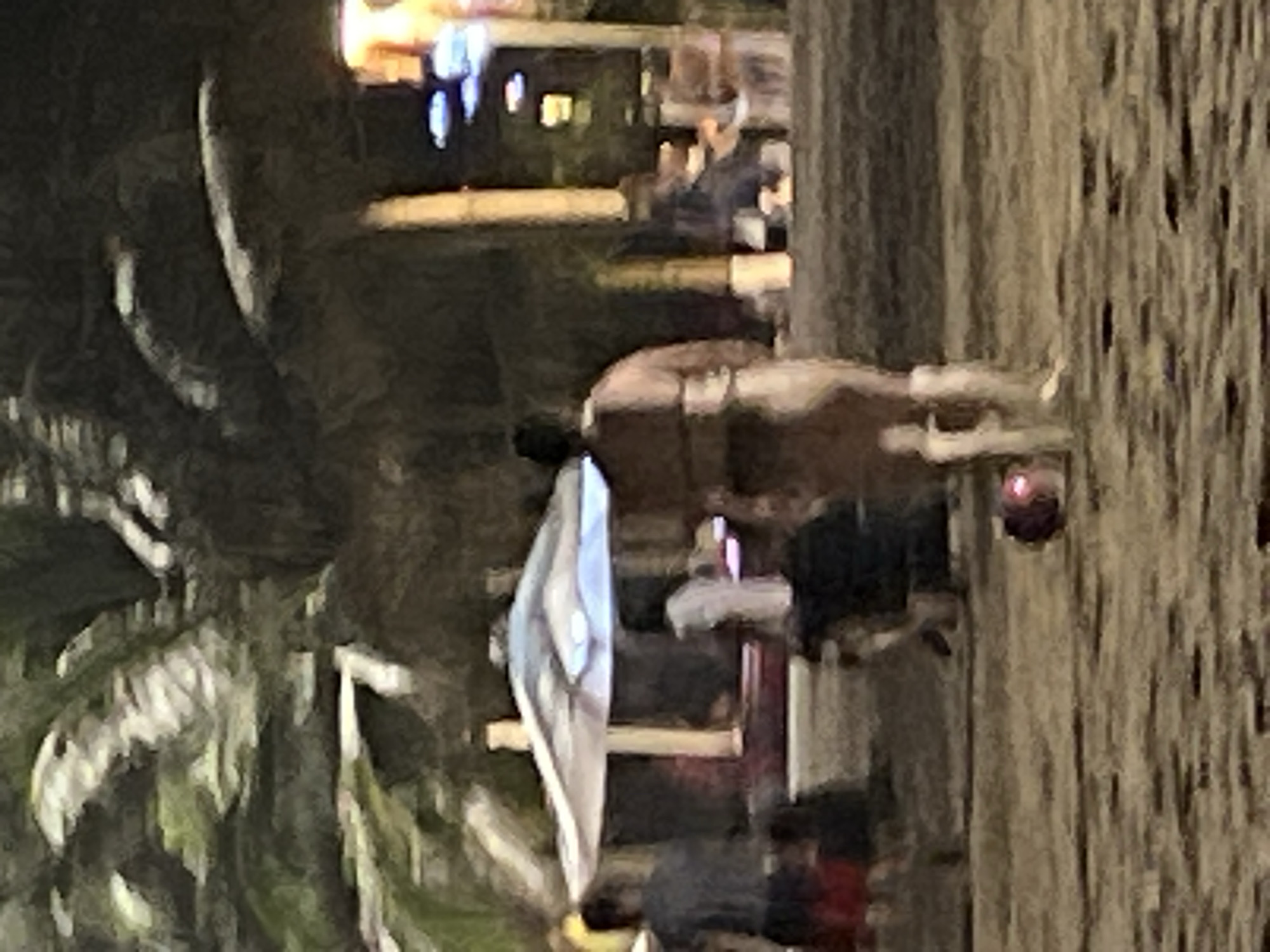 The image depicts a nighttime scene on a sandy beach, likely in Nha Trang, Vietnam, with palm trees and illuminated structures in the background. In the foreground, a person is walking away from the camera, wearing light-colored footwear and a light-colored garment. To the left, a person in a dark shirt and red shorts is partially visible, walking away from the camera. Further to the left, an umbrella shades a seating area. In the middle ground, another person is standing. The ground is covered in sand and small pebbles, with a red, spherical object on the sand to the left of the foreground figure. In the background, there are lights and structures, suggesting a developed area, possibly a promenade or entertainment zone.