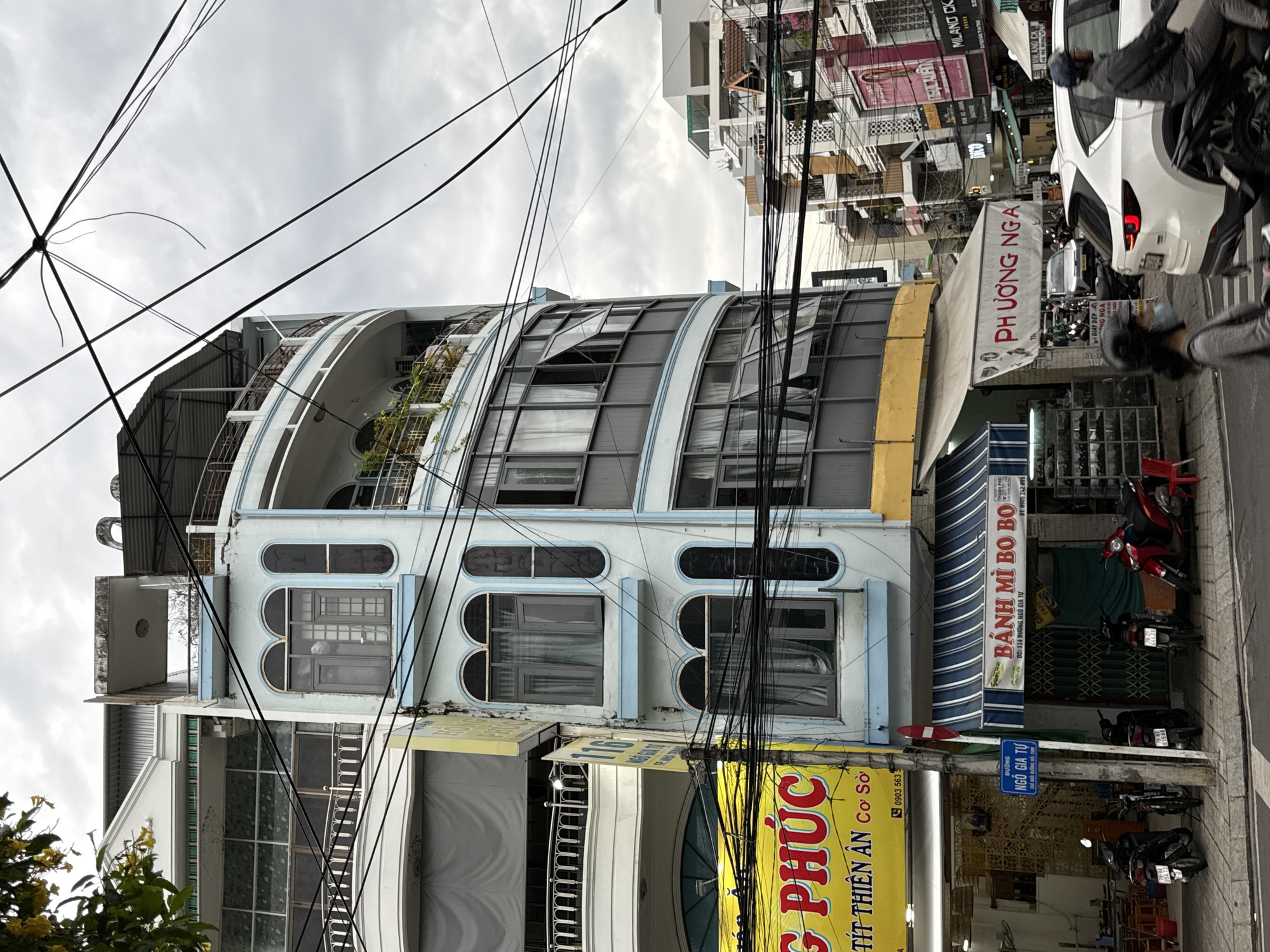 The image depicts a street scene in Nha Trang, Vietnam, characterized by a dense urban environment with multi-story buildings, overhead electrical wires, and a cloudy sky. The foreground features a light blue, curved building with several floors, adorned with arched windows and balconies. A yellow sign with Vietnamese text is visible on the left side of the building, and a blue and white striped awning extends from the ground floor. Several motorcycles are parked along the sidewalk in front of shops. Further down the street, other buildings with various architectural styles and signs are visible. Two people are seen on motorcycles, one in the foreground and another in the mid-ground. The overall impression is one of a bustling city street under an overcast sky.