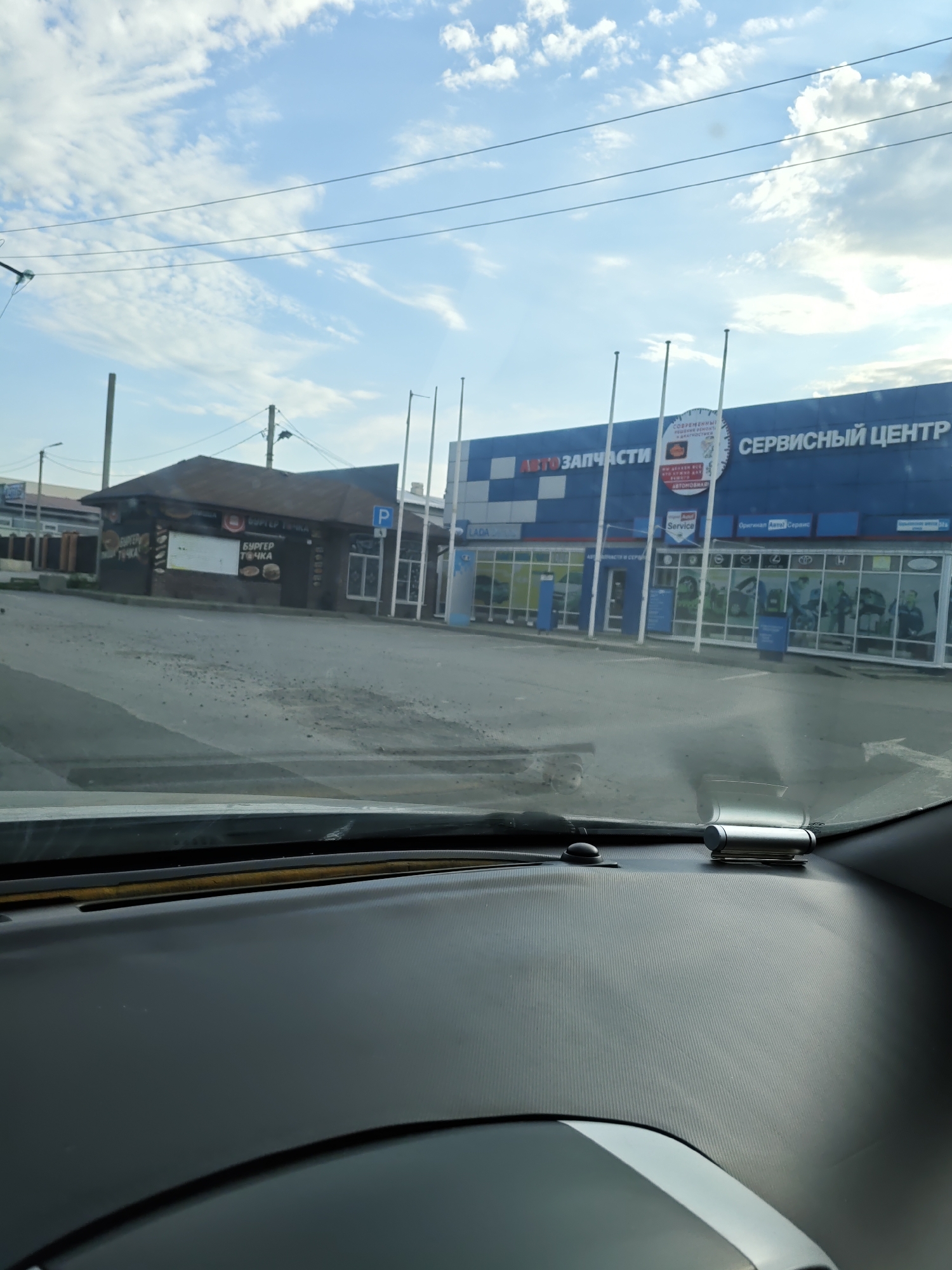 The image was taken from inside a vehicle, looking out through the windshield. The exterior scene shows a street in Novocherkassk, Russia, with a blue and white building in the background that appears to be an auto service center with a sign reading "АВТОЗАПЧАСТИ" (Auto Parts) and "СЕРВИСНЫЙ ЦЕНТР" (Service Center). To the left of this building is a smaller, dark-colored structure with a brown roof, which seems to be a fast-food establishment with signs displaying "БУРГЕР ТОЧКА" (Burger Point). The street in front of the buildings is paved and appears somewhat weathered. Overhead, telephone or power lines are visible against a sky with scattered white clouds. The interior of the vehicle shows the dashboard in the foreground, with a silver device attached to the windshield.