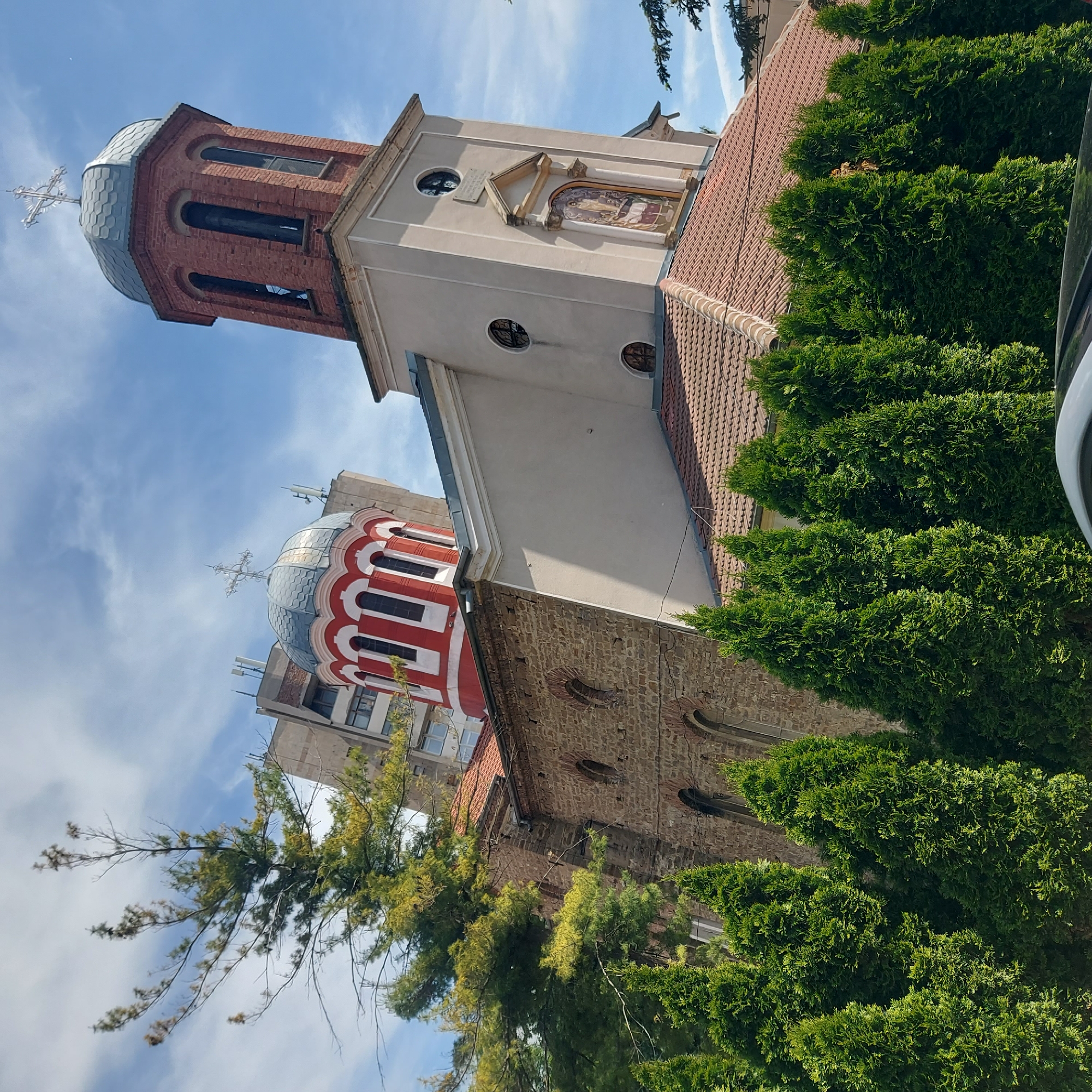 The image shows the Holy Trinity Church in Gorna Oryahovitsa, Bulgaria. The church has a tower with a brick top and a smaller tower with a gray and red dome. The main building is made of brick and stone, with a light-colored upper section. The church is surrounded by greenery, including trees and bushes. The sky is blue with some clouds.