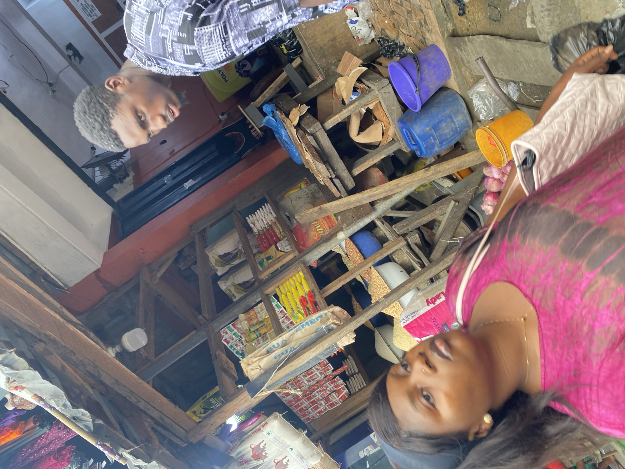 The image shows an indoor scene in Lekki, Nigeria. Two individuals are visible: a person wearing a patterned black and white shirt and another wearing a pink top. They appear to be standing within what seems to be a shop, judging from the shelves stocked with goods and various buckets and containers. There are sacks labeled "Lady" and "BIG BULL" indicating rice, as well as items labeled "BUA" and "LION". The shop's structure has exposed wooden beams, and a lightbulb is visible on the ceiling.