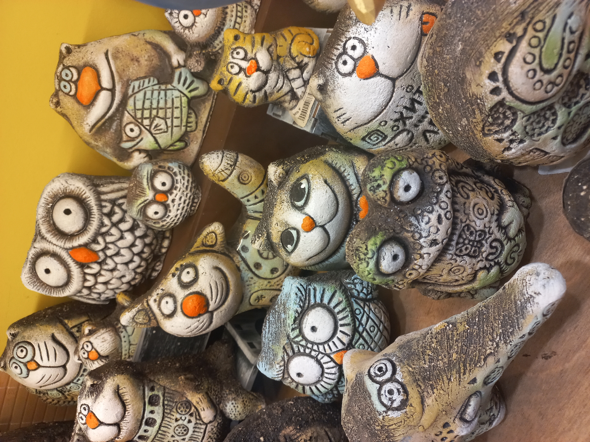 The image shows a high-angle, close-up view of a collection of ceramic animal figurines, likely for sale. The figurines are densely packed together, obscuring details of their arrangement. Predominantly, the figurines depict stylized owls and cats. Other animals such as a tiger and a cat carrying a fish are also present. The figurines are decorated with various patterns and painted in muted tones of gray, white, yellow, and orange. A shelf is visible at the back and at the right of the scene. A yellow wall is also visible. A price sticker is visible on one of the figurines. There are no people in the scene. The city and country are not clear from the image.