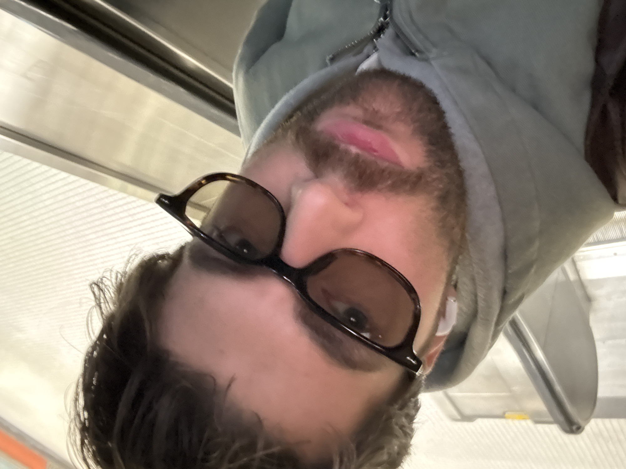 The media file is an image rotated 90 degrees counter-clockwise, capturing a close-up selfie of a man. The man is looking upwards towards the camera, with a neutral expression. He has dark hair, a beard, and is wearing tortoiseshell-framed sunglasses with dark lenses. A white earbud is visible in his right ear. He is dressed in a light grey or light blue hooded jacket, with a zipper visible at the neckline. A reddish mark or sore is visible on his upper lip, slightly to the right of his philtrum.
The background indicates an indoor public environment, likely a transit station or a building featuring a moving walkway or escalator, which aligns with the provided context of Montreal, Canada. Metallic structures, including what appears to be a handrail and the ridged surface of an escalator step, are visible. The scene is illuminated by artificial indoor lighting. No visible text is present in the image.